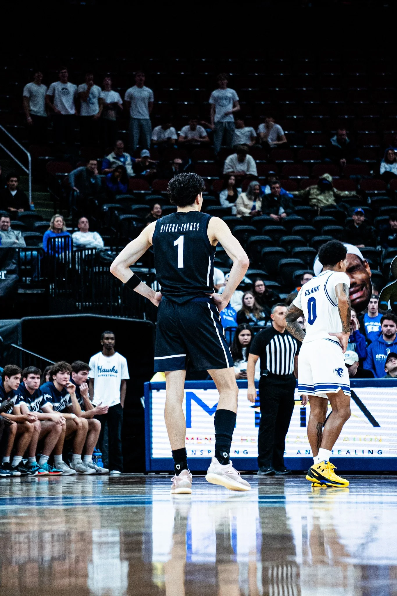 A basketball game scene with players on the court, including a player with jersey number 1 and another with jersey number 0. The players appear to be taking a break, talking with each other and the coach. Spectators are seated in the background, watc