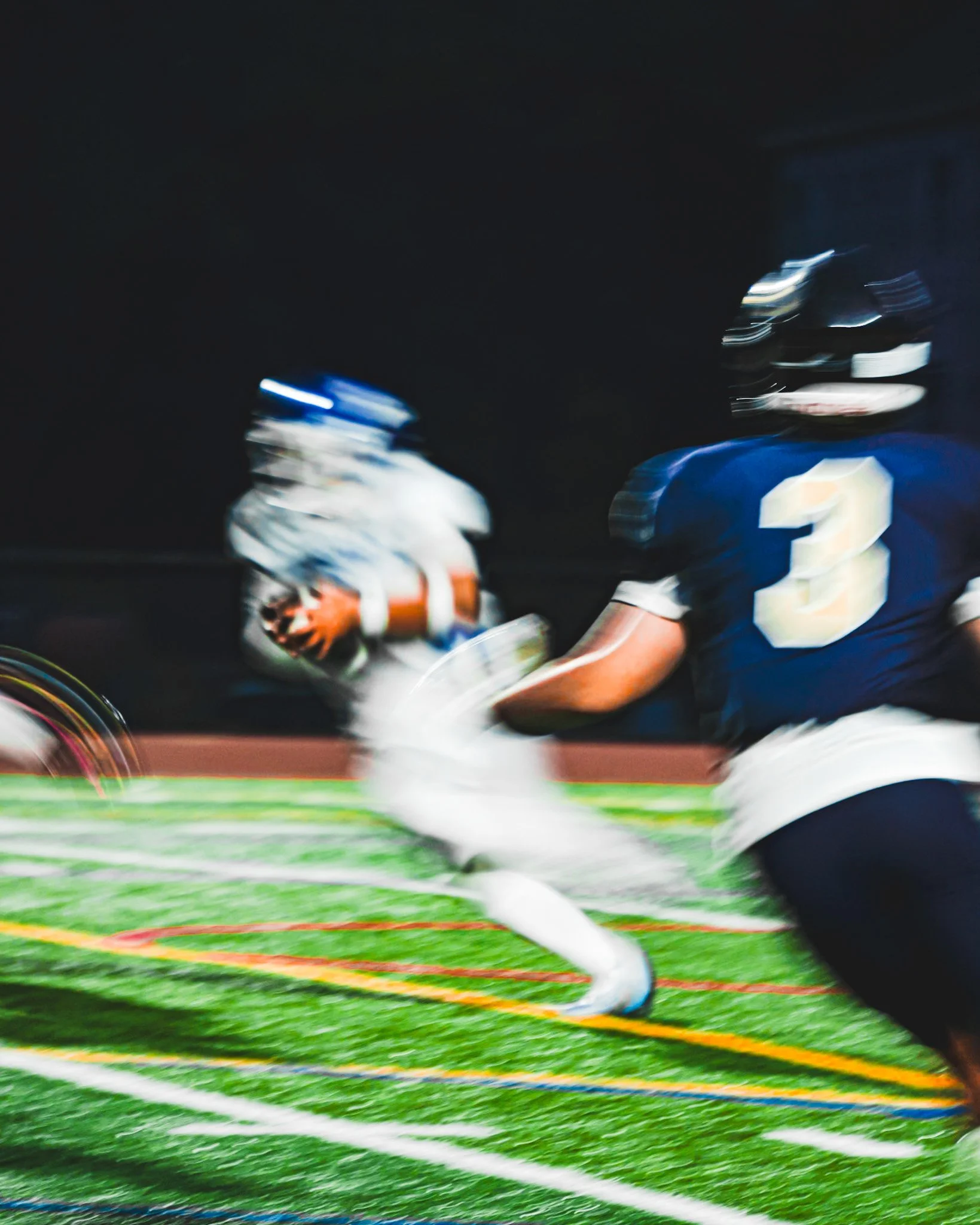 Blurred image of two football players on the field during a game, one wearing a white uniform with the number 10, the other wearing a blue uniform with the number 3, with colorful lines on the bright green field.