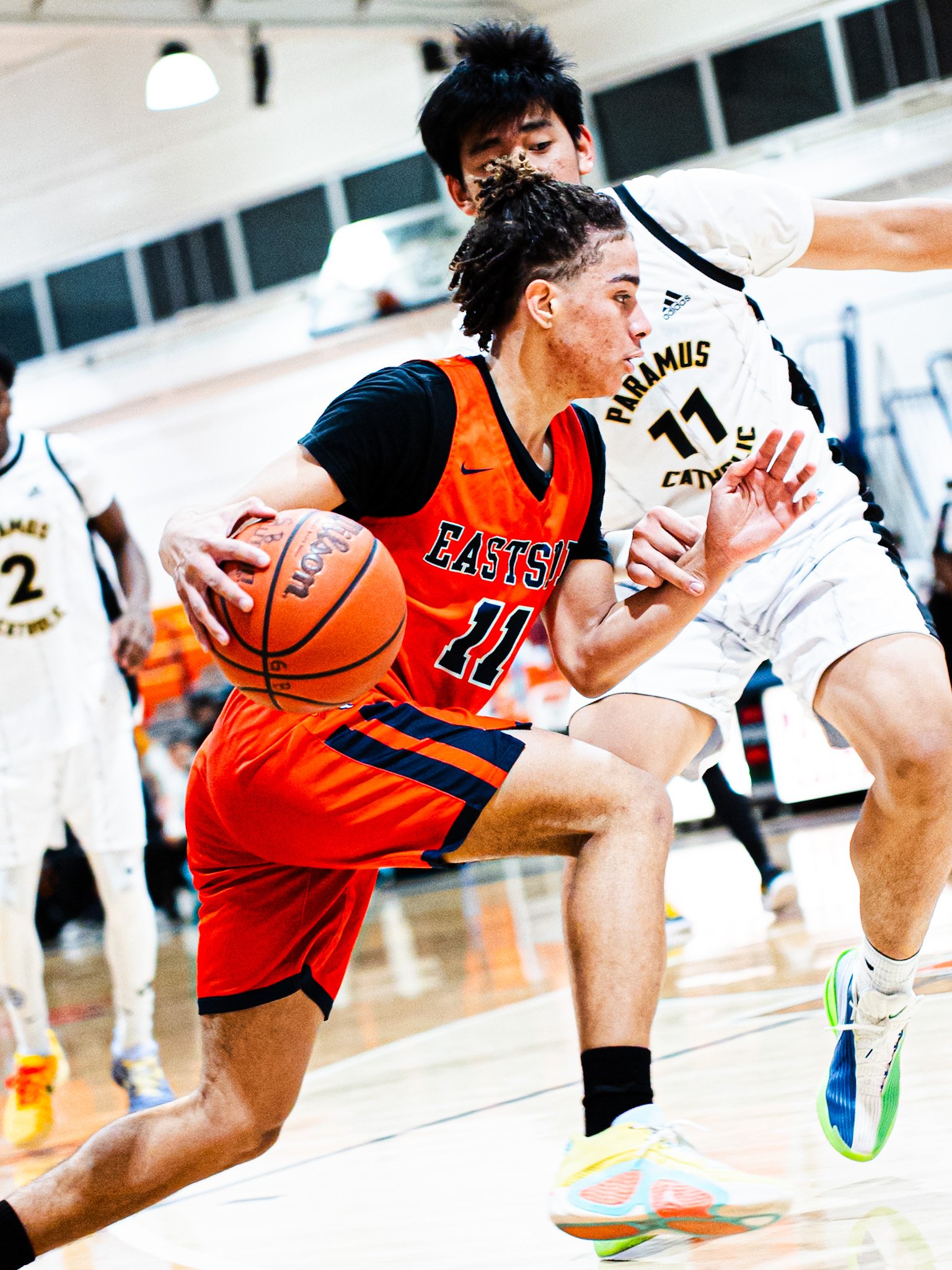 Two male basketball players in action during a game. The player in the orange and black uniform is holding a basketball and is leaning forward, while the player in the white uniform attempts to block or defend.