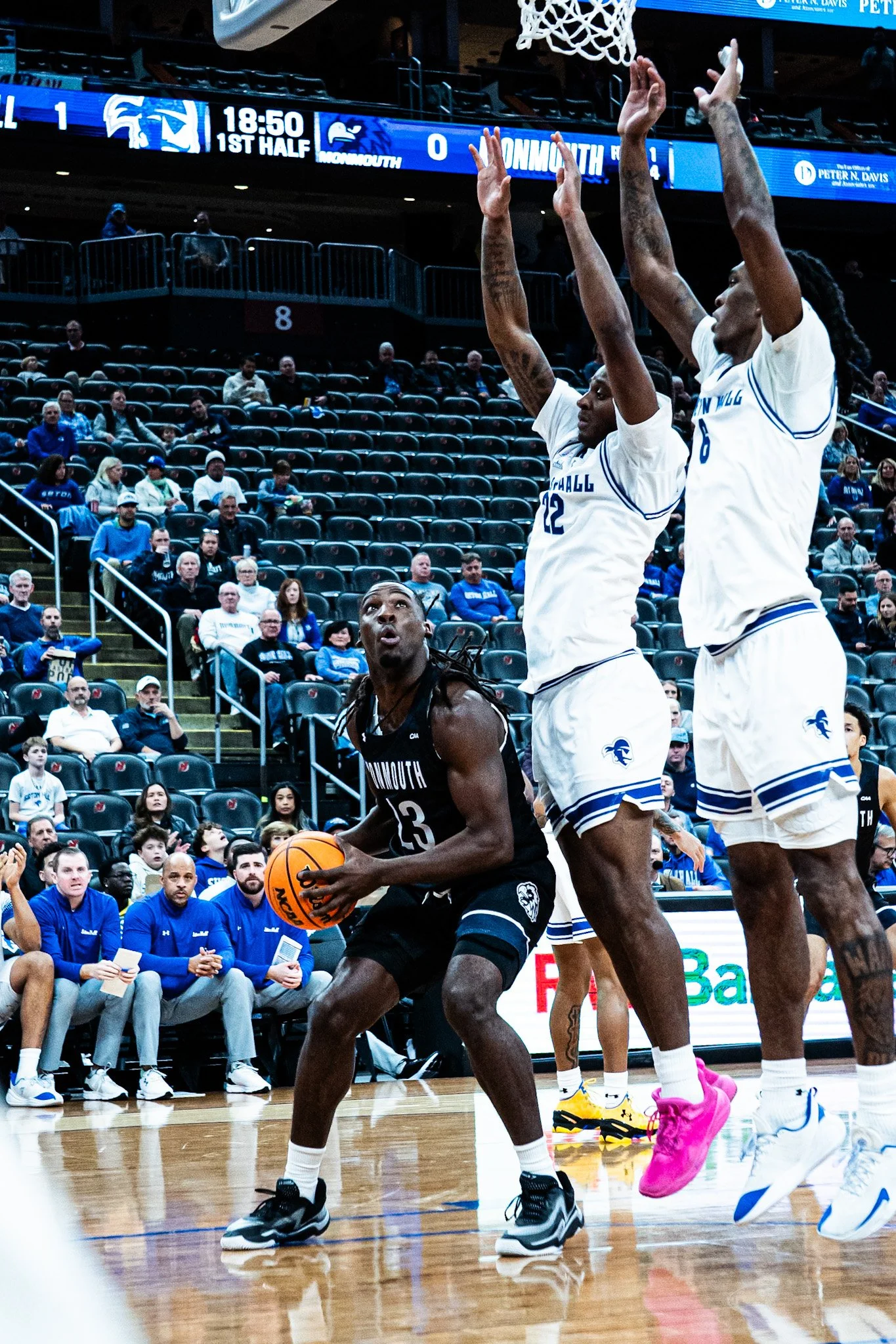 Two basketball players in white jerseys are jumping with their arms raised as a player in a black jersey dribbles a basketball on a court. Spectators and team officials are seated and watching in the background.