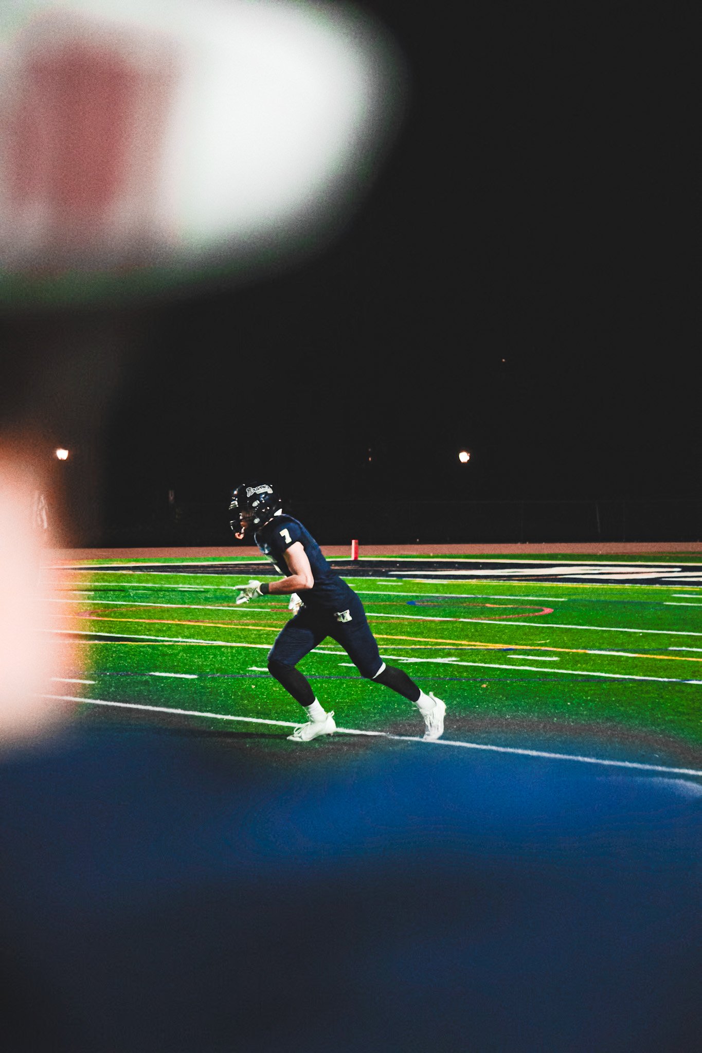 A football player running on a field at night, wearing a black jersey with the number 7, a helmet, and cleats, with stadium lights overhead and a track around the field.