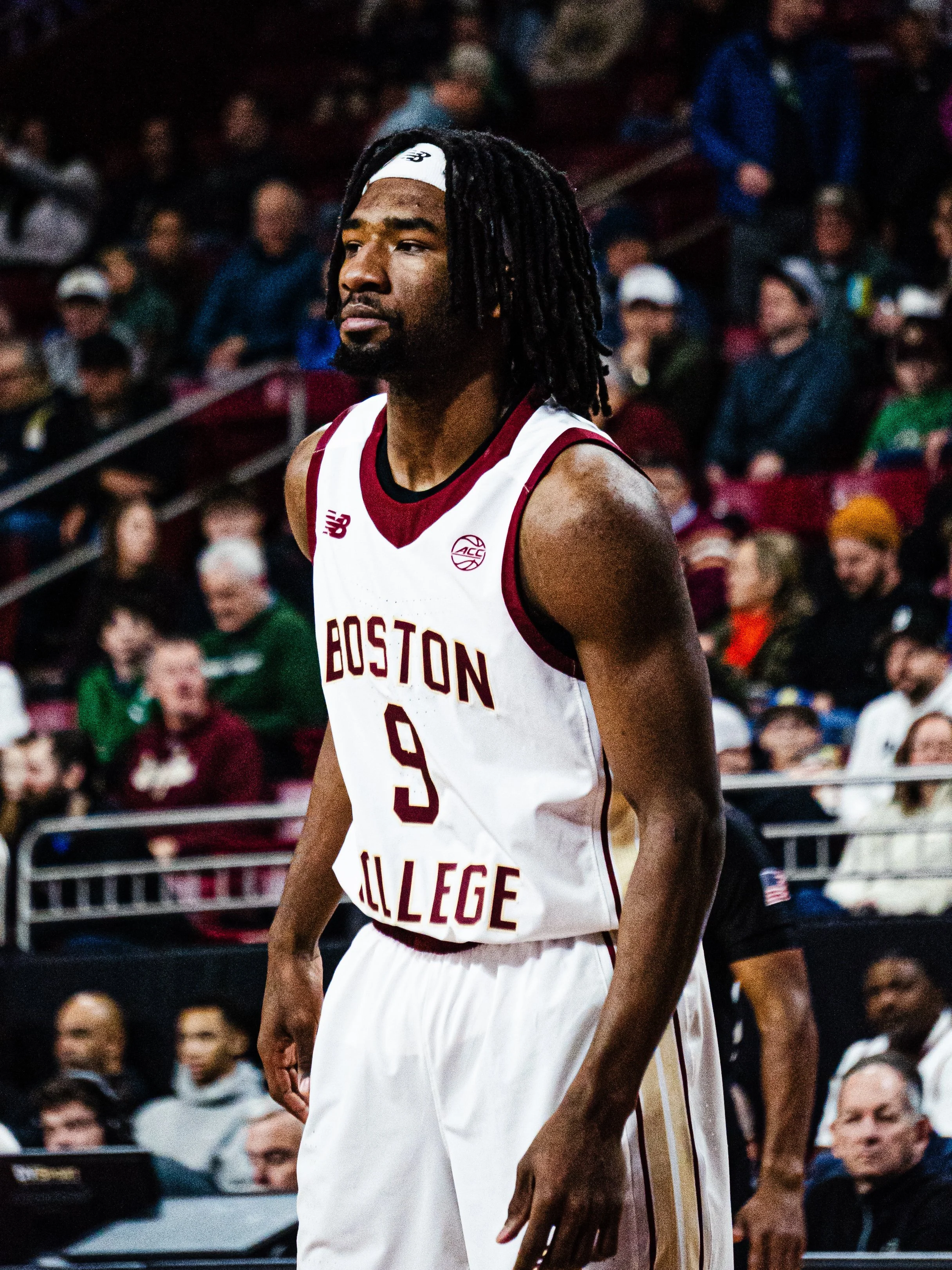A basketball player from Boston College stands on the court with a crowd in the background.
