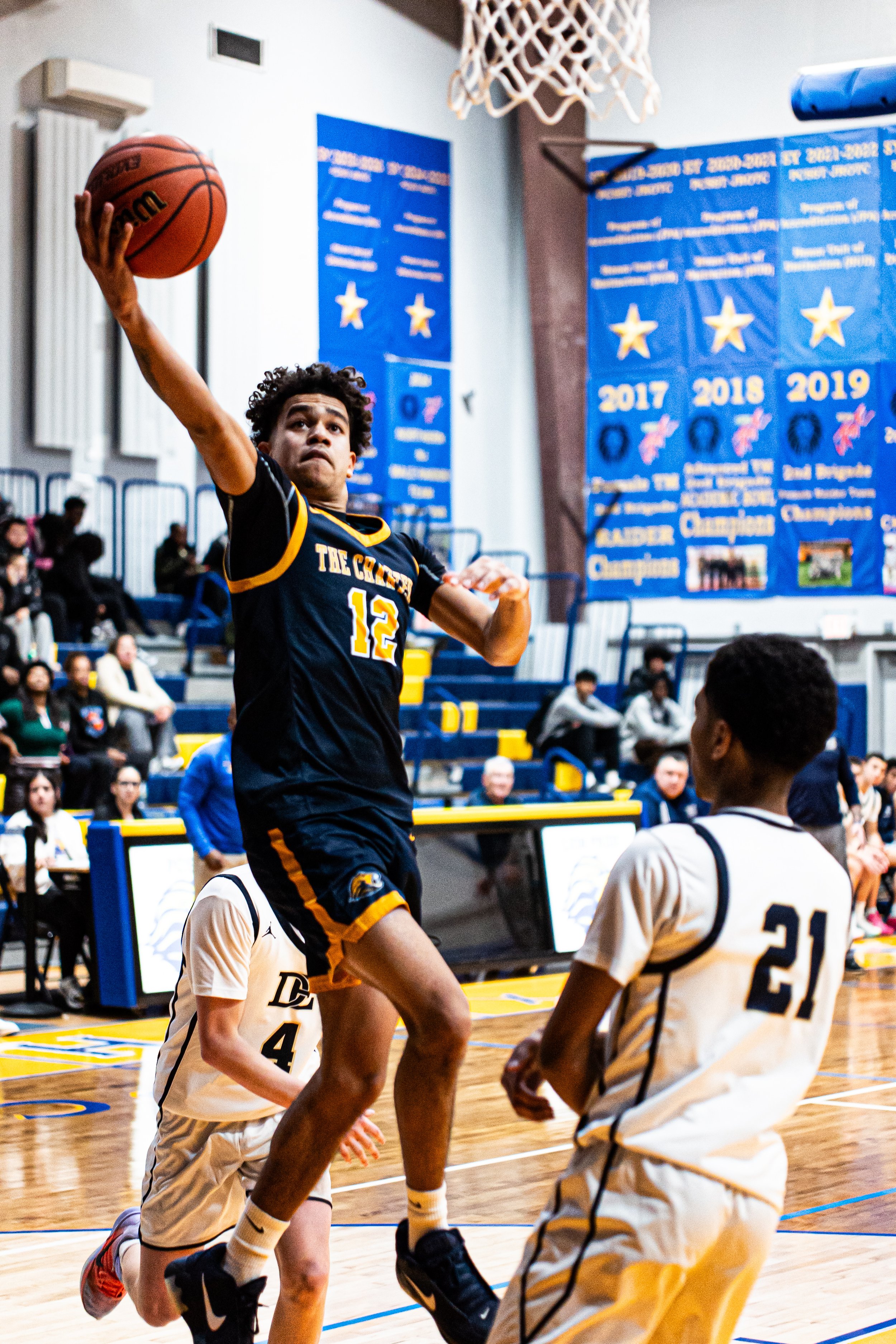 A basketball player in a navy blue and gold uniform jumps to layup during a game in a gymnasium. Opposing players in white uniforms with black trim are defending, with one player kneeling and another standing nearby. Spectators watch from bleachers. 