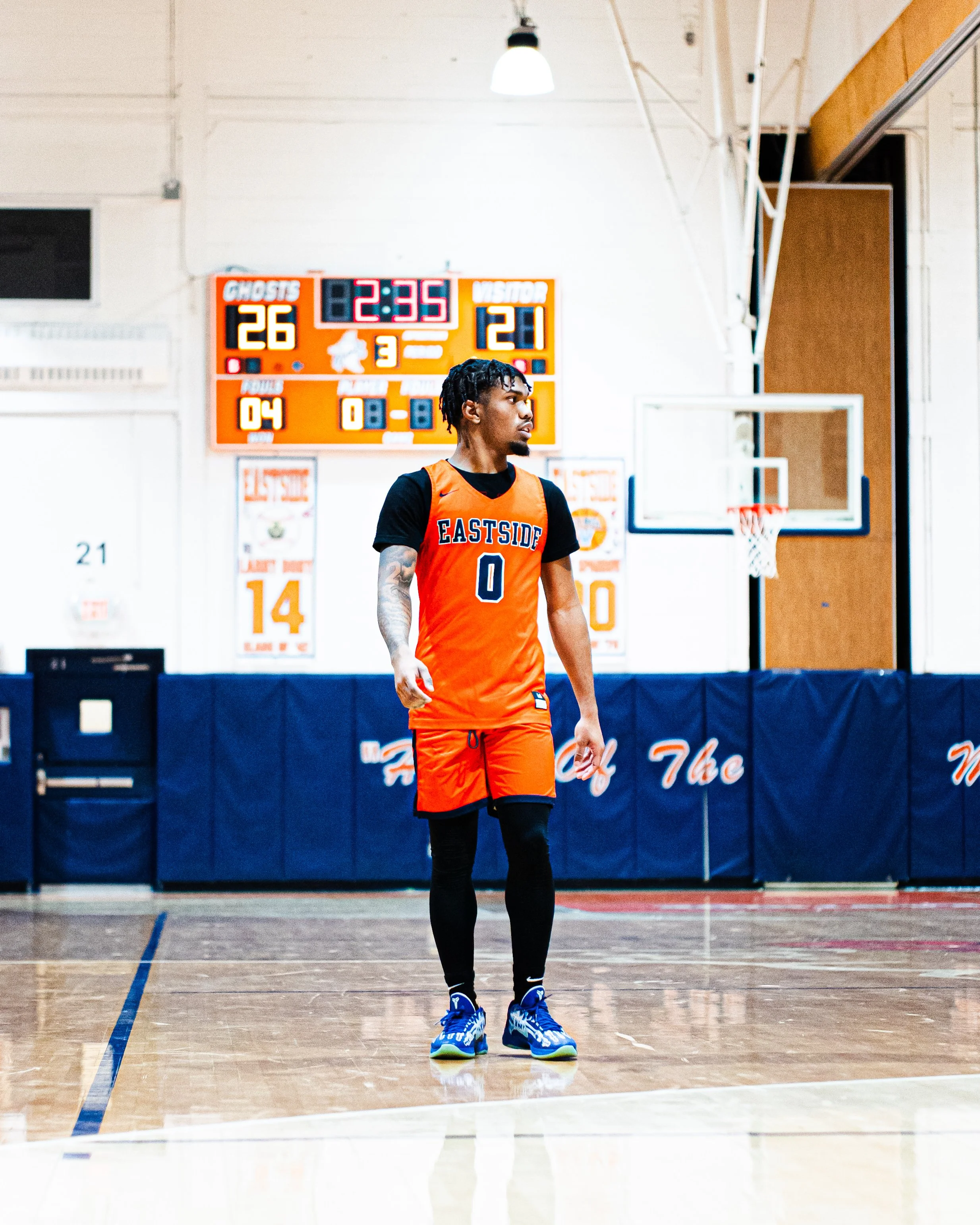 A basketball player in an orange jersey with 'EASTSIDE' and the number 0, standing on an indoor basketball court, with a scoreboard in the background showing 26-21 in favor of the ghosts, and 2 minutes and 35 seconds remaining.