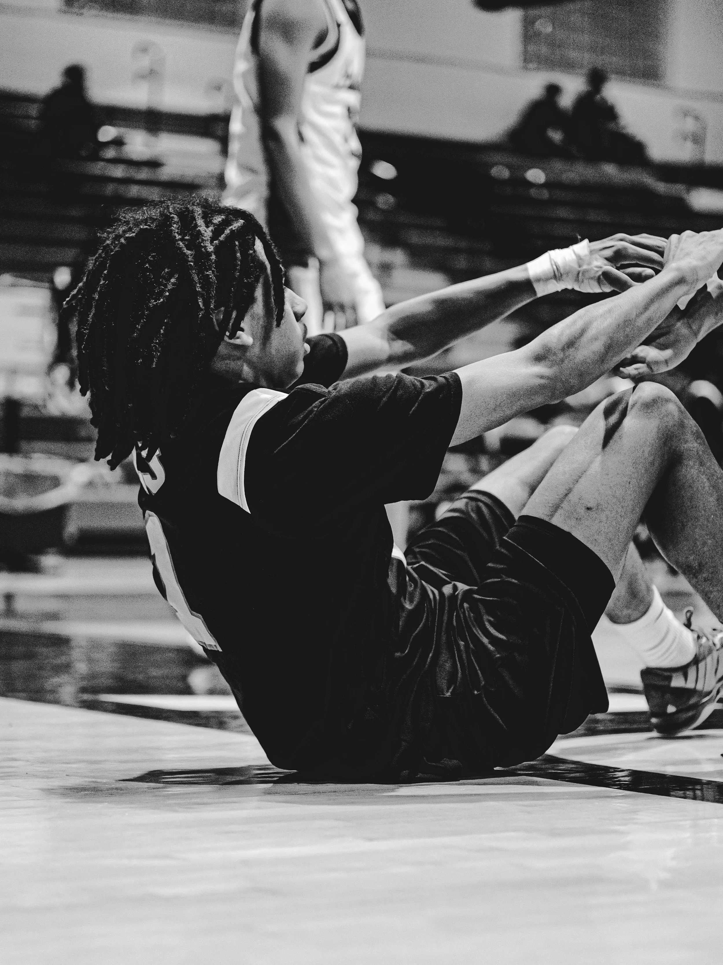 A person with dreadlocks falling backwards on a gymnasium floor, appearing to have fallen or is in a stretching or exercise position, with other people sitting on benches in the background.