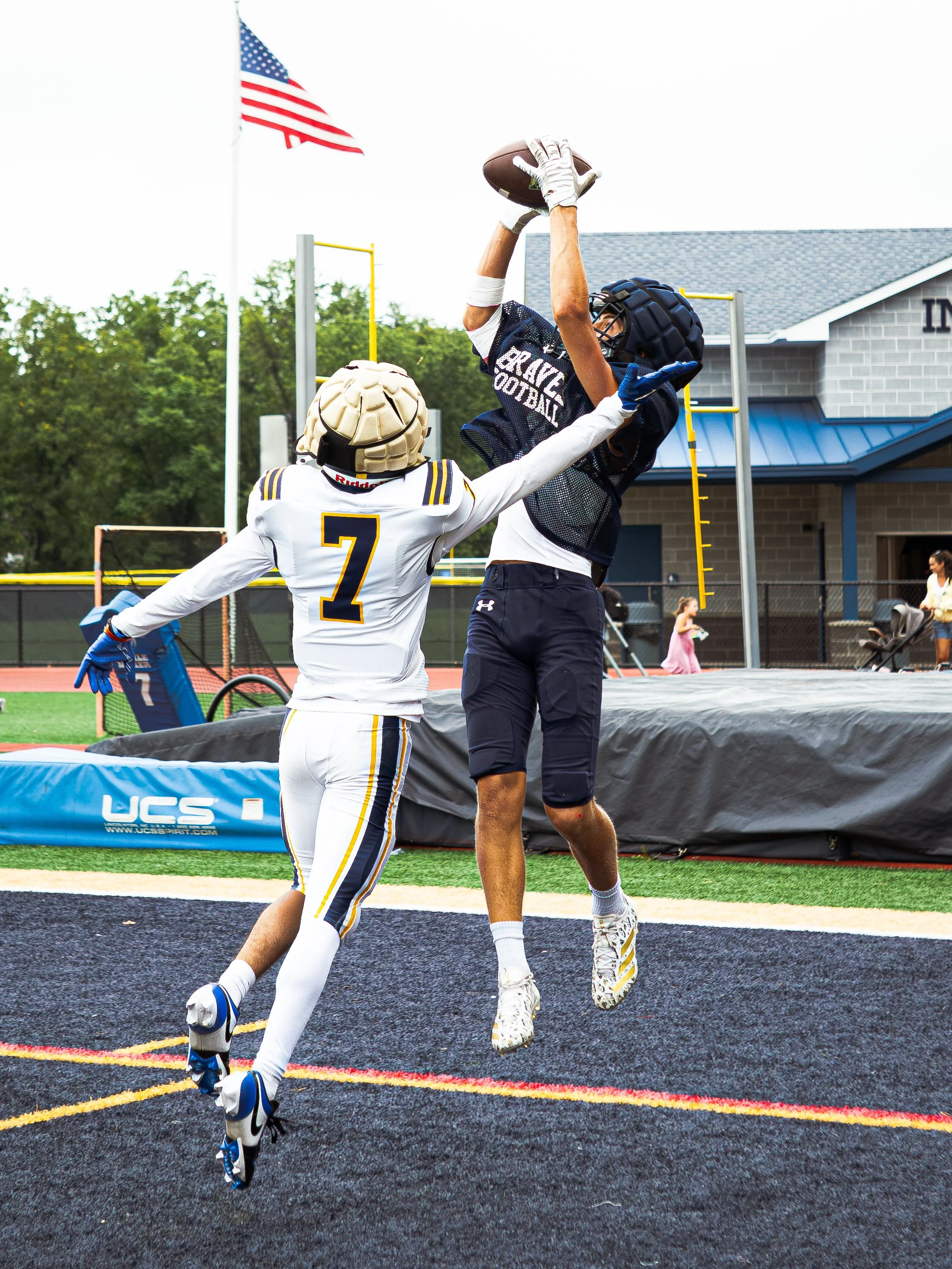 Two football players are jumping to catch a football near the end zone on a football field. One player wears a black helmet and navy jersey, the other wears a tan helmet and white jersey with the number 7. There is an American flag in the background.