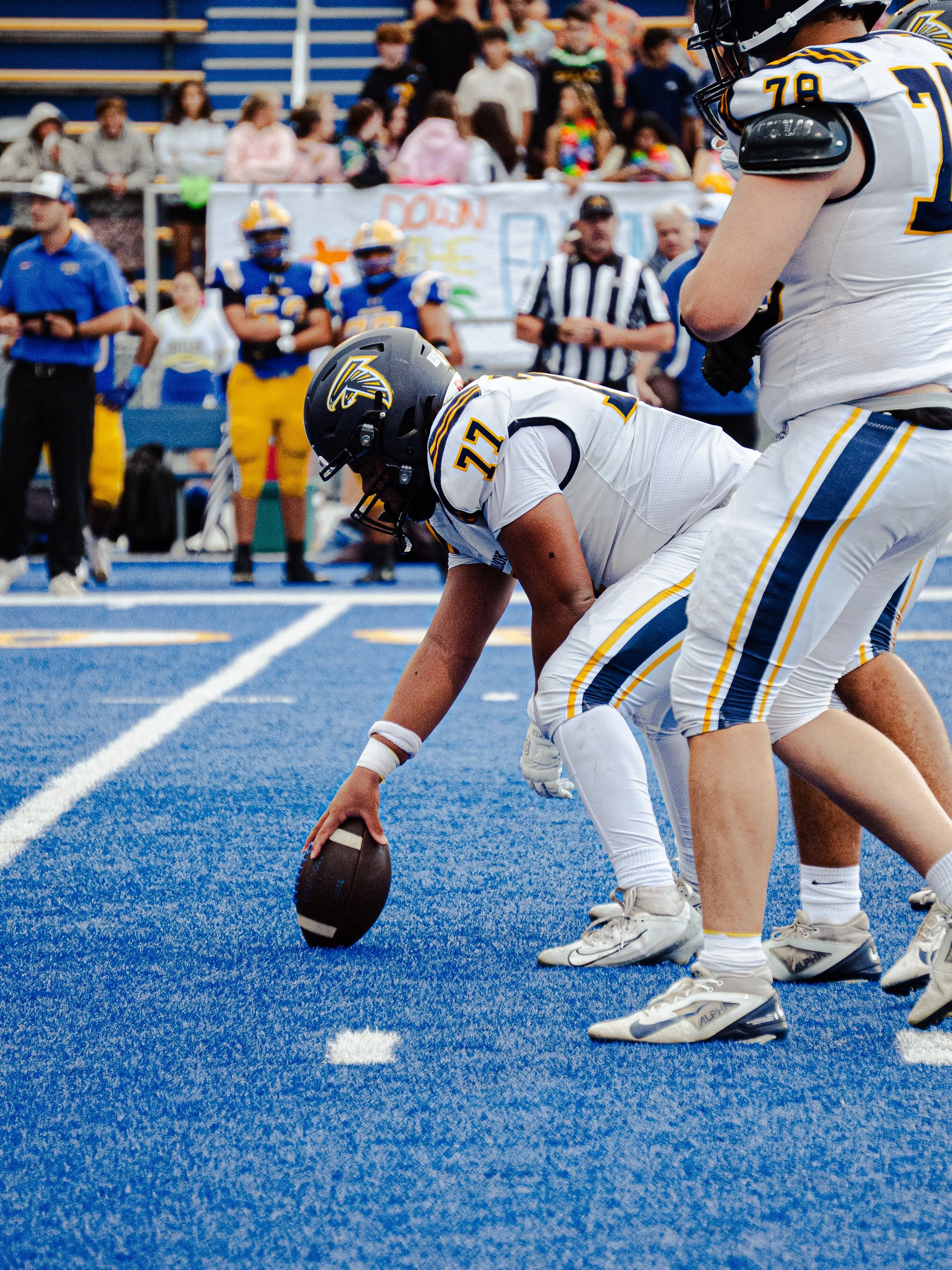 American football players in white uniforms with yellow and blue accents lining up on a bright blue field before a play, with spectators and team members in the background.