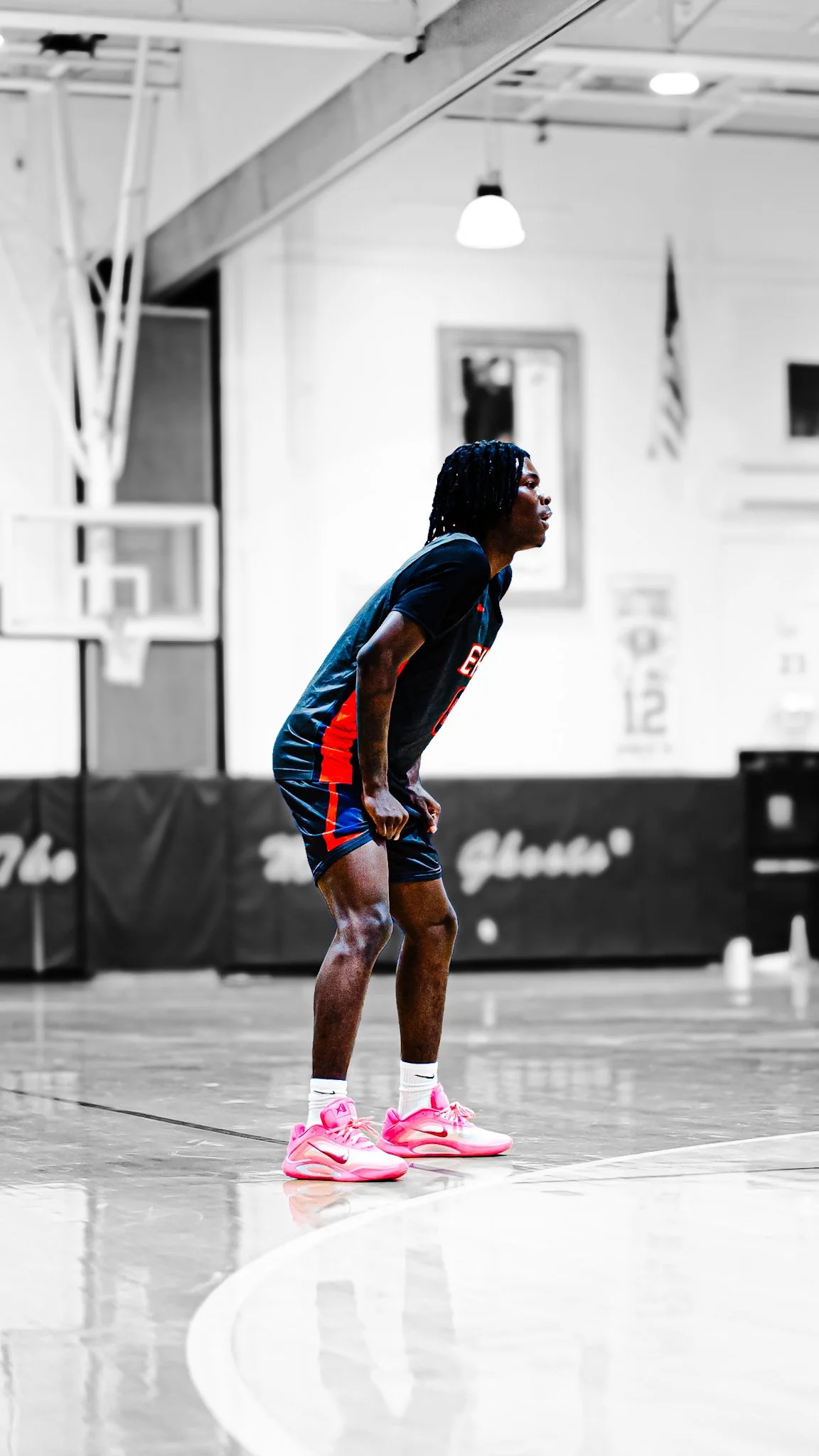 A basketball player, wearing a black jersey and pink sneakers, is standing on a basketball court in an indoor gym, bent over with hands on her knees, appearing to be tired or resting.