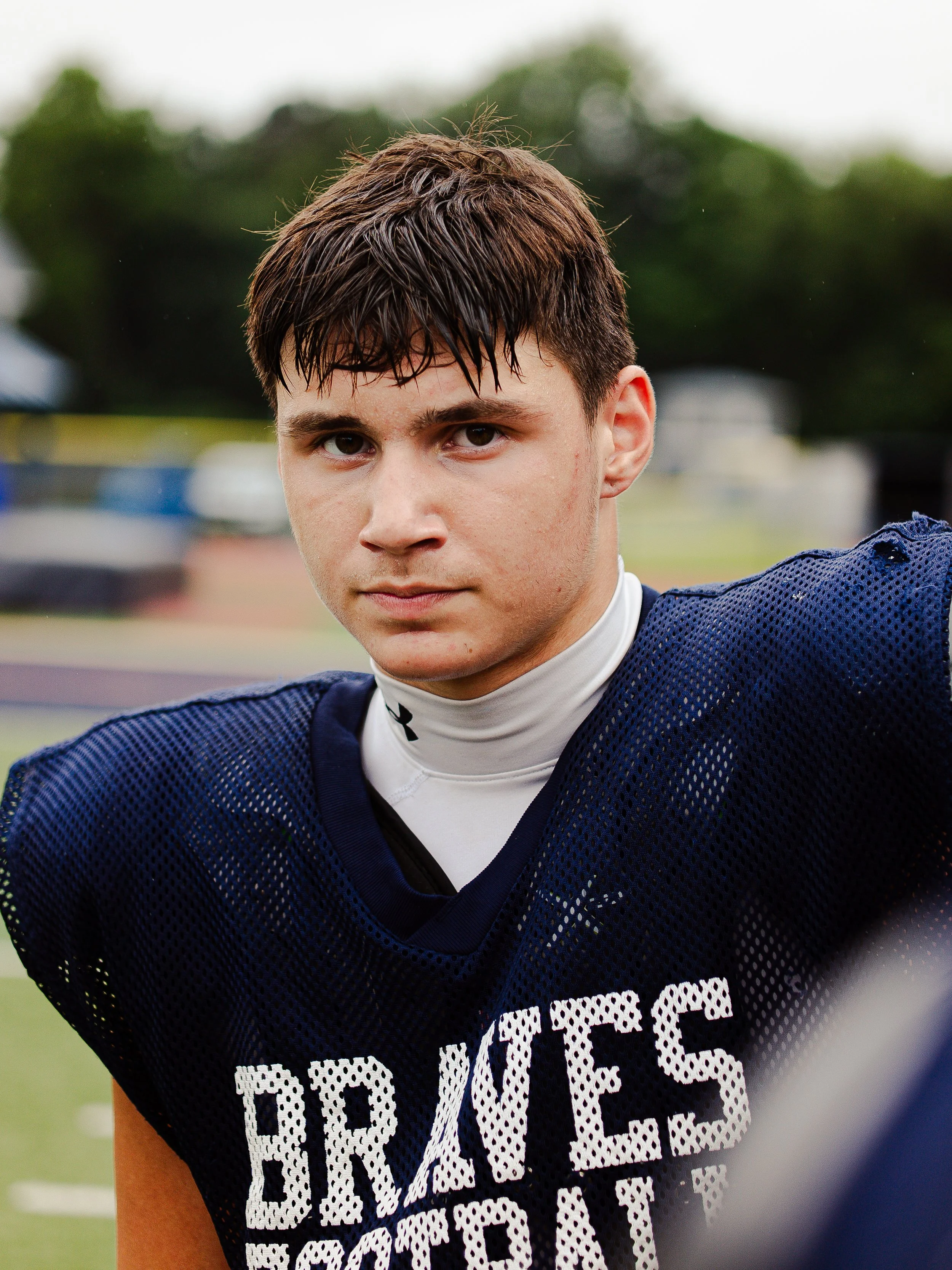 Close-up of a young male football player wearing a dark blue practice jersey with white lettering, standing on a field with trees in the background.