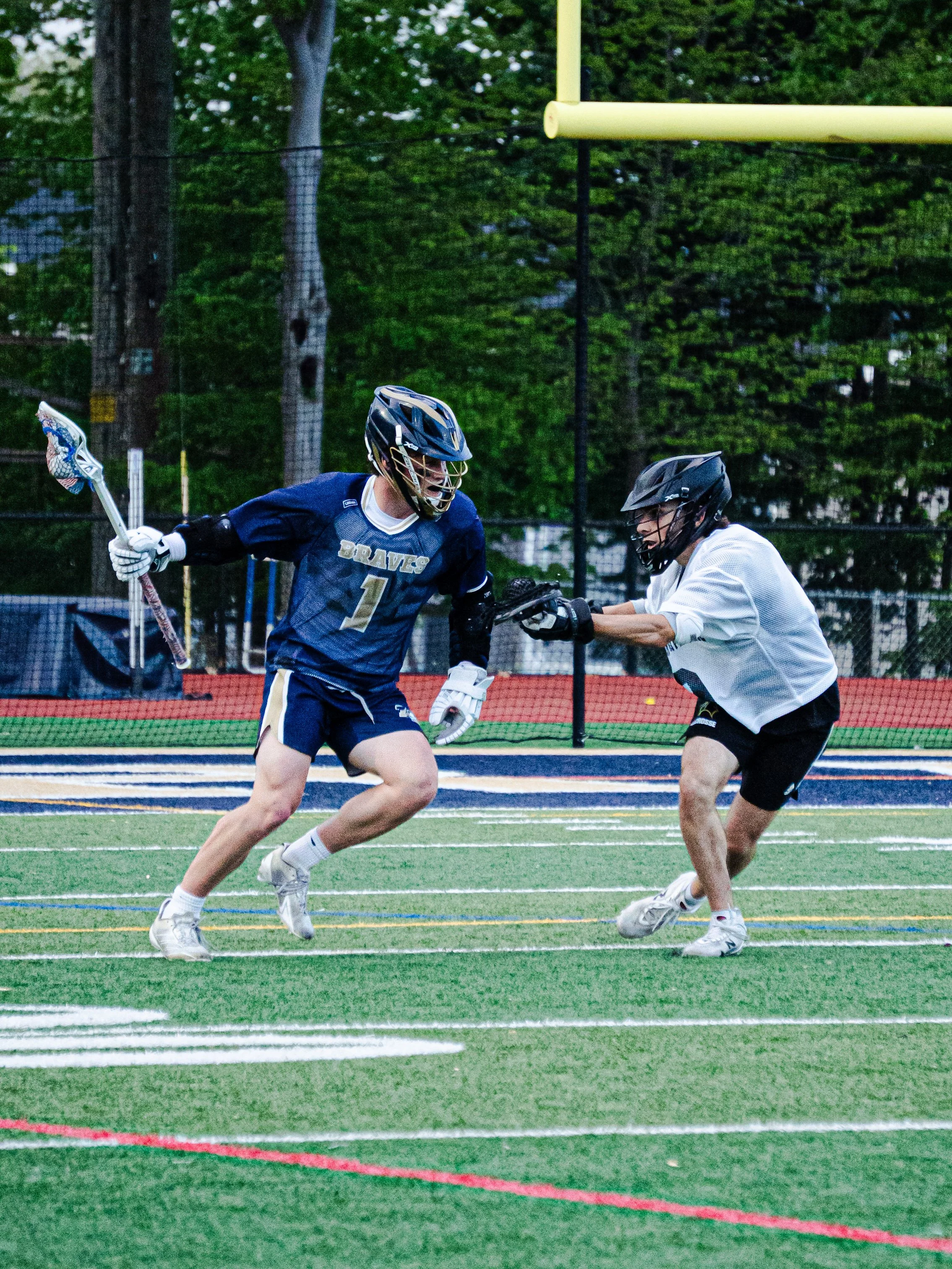 Two boys playing lacrosse on a sports field during a game, wearing helmets and sports uniforms, with a goal post visible in the background.