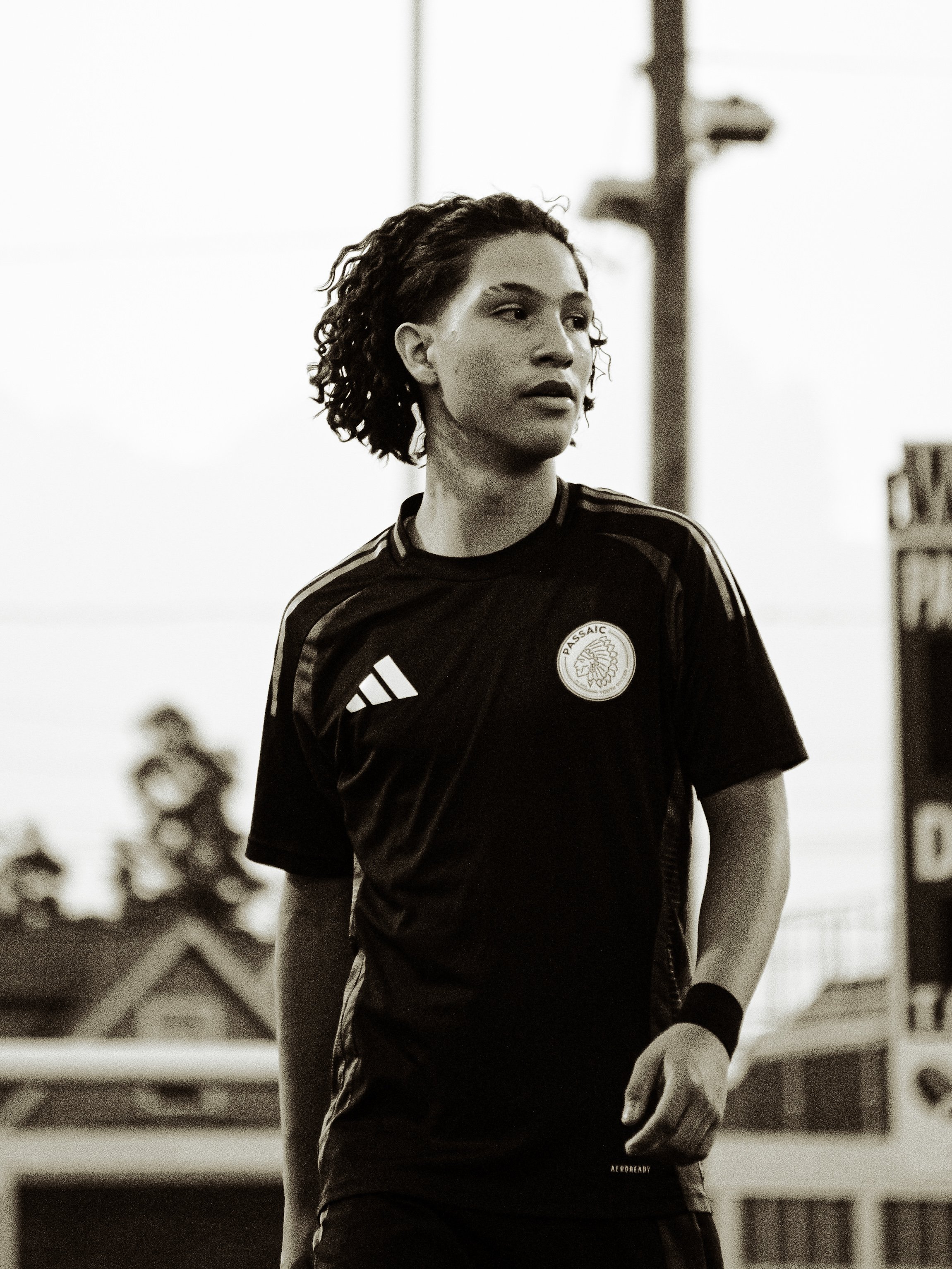 Young woman with curly hair wearing a sports jersey, standing outdoors with a blurred background of trees and a building.