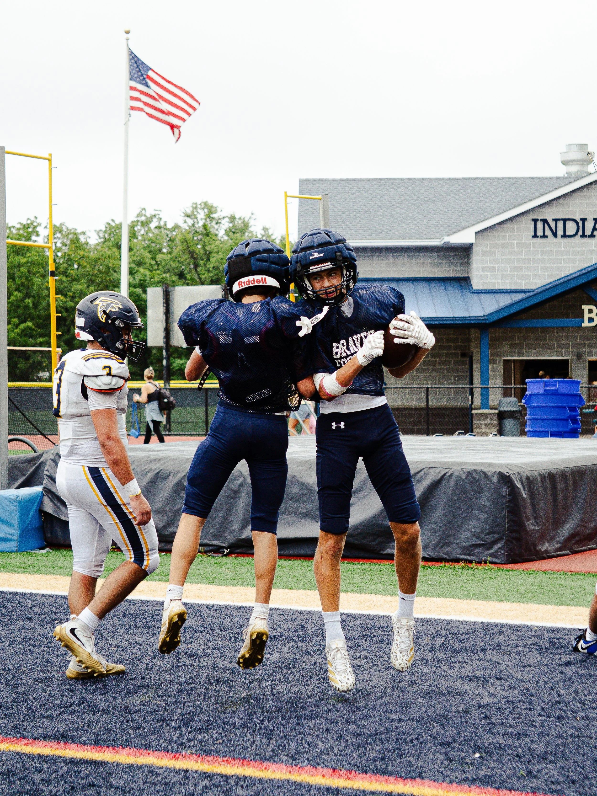 High school football players celebrating on the field, with one player holding a football, others jumping, and a coach or teammate in the background.