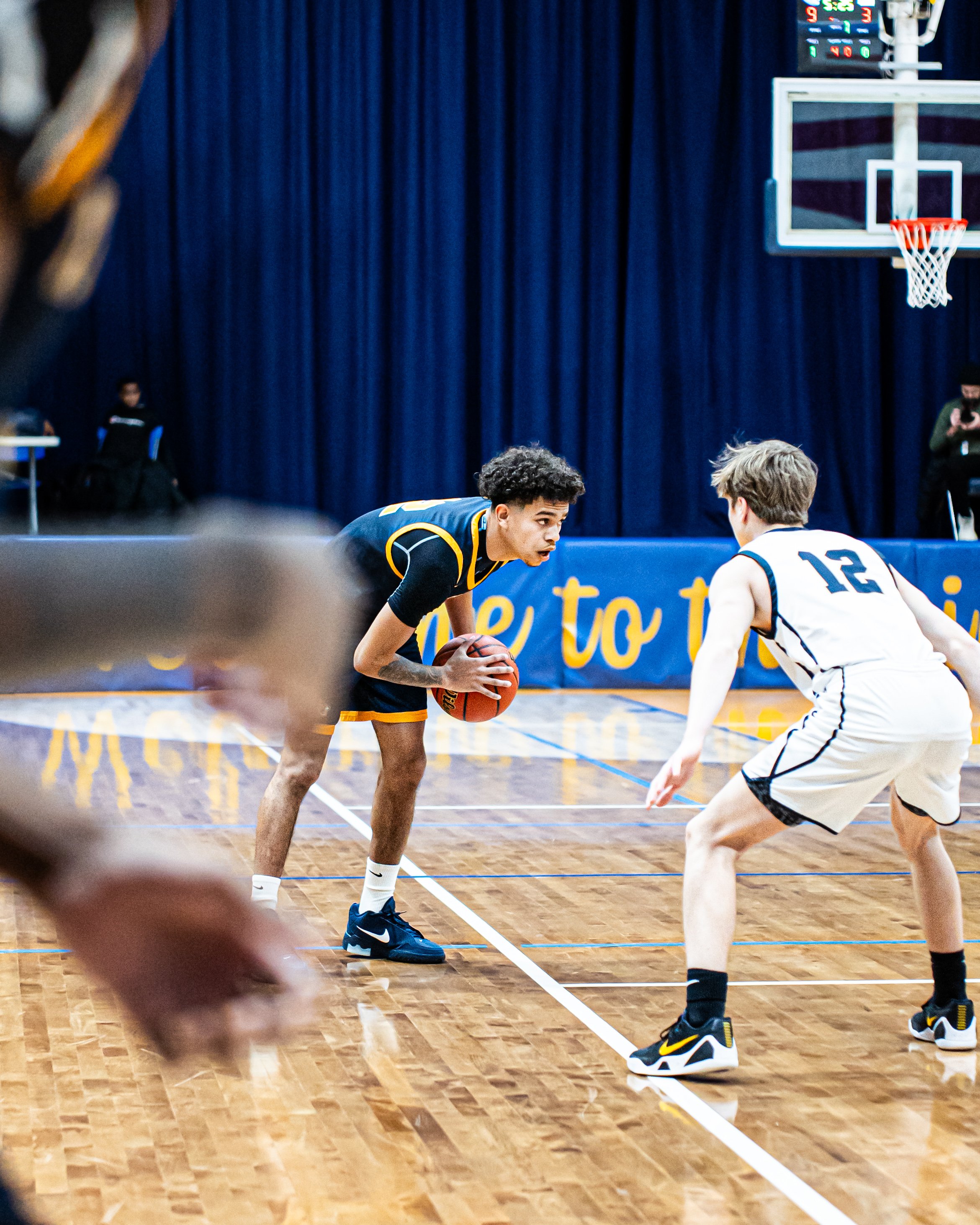 Two basketball players, one in a dark uniform and the other in a white uniform, are facing each other on the court during a game. The player in the dark uniform is holding a basketball, and the game is taking place in an indoor gym with a blue curtai