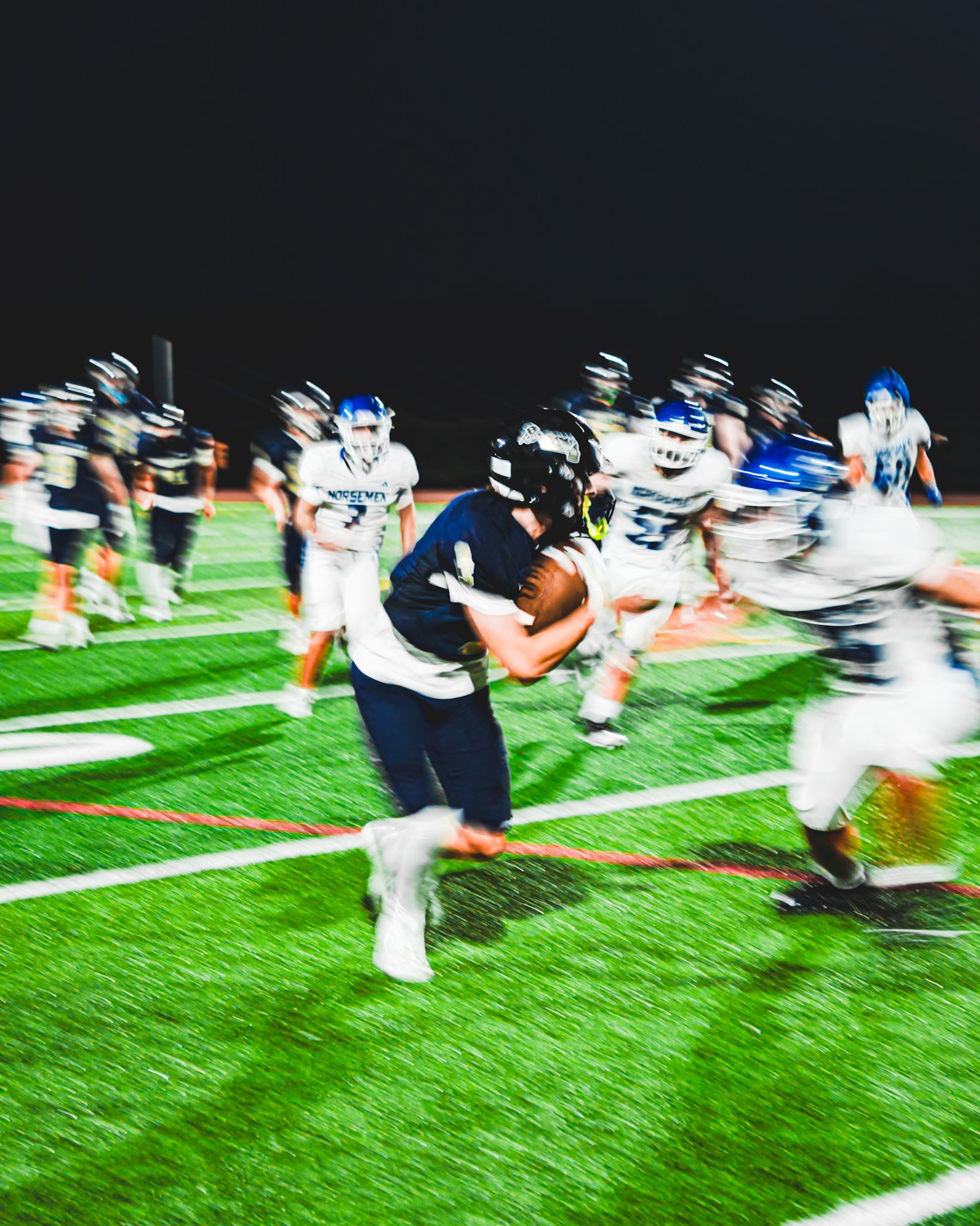 Football players on a field during a night game, with some players in white uniforms and others in dark uniforms, and a referee in black in the foreground.