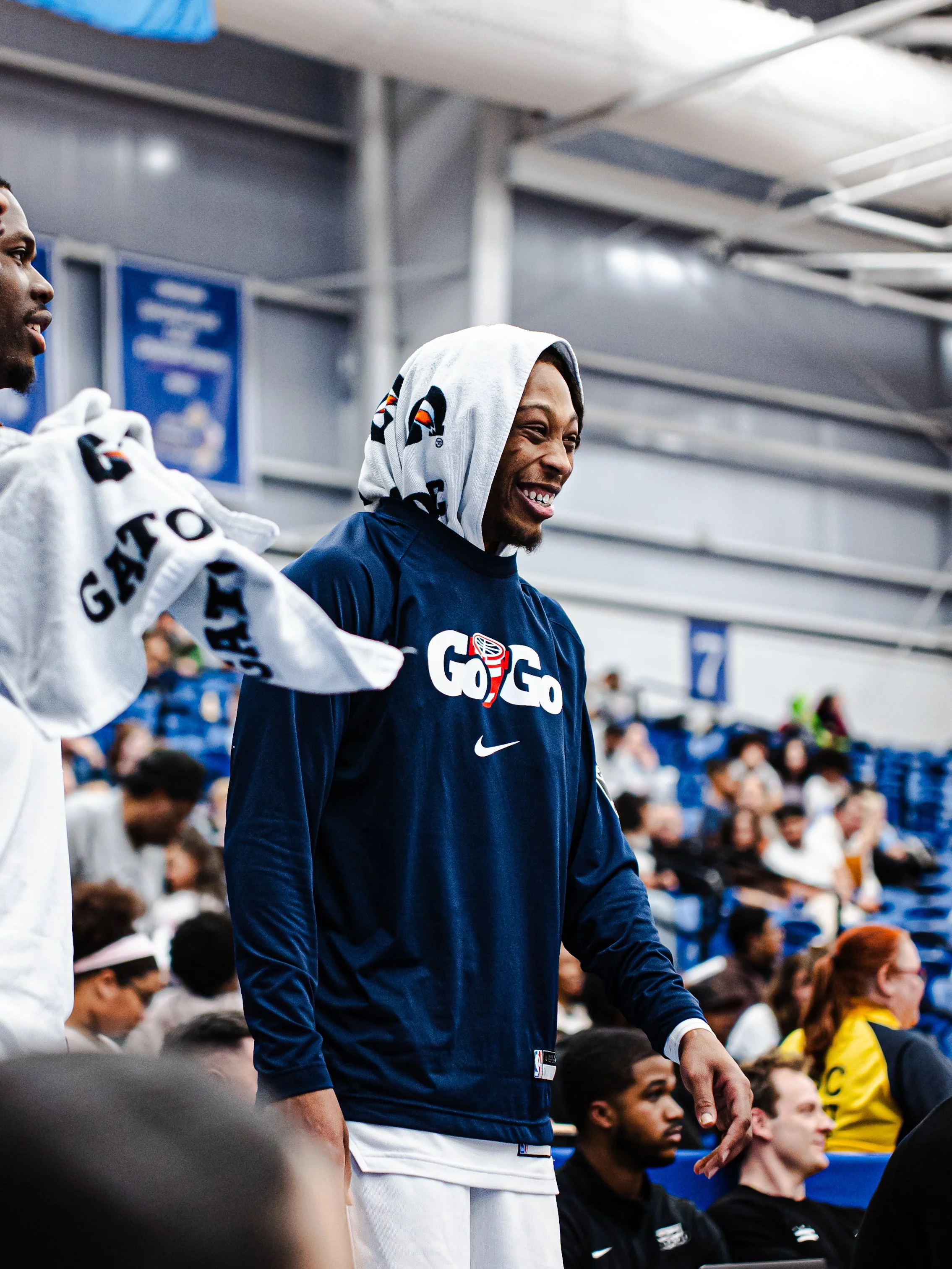 Group of people, likely athletes, sitting in a stadium or sports arena, with some standing. One person in the foreground is smiling, wearing a hoodie with a towel on their head, and has a navy blue long-sleeve shirt with the logo 'GOG' and a white Ni