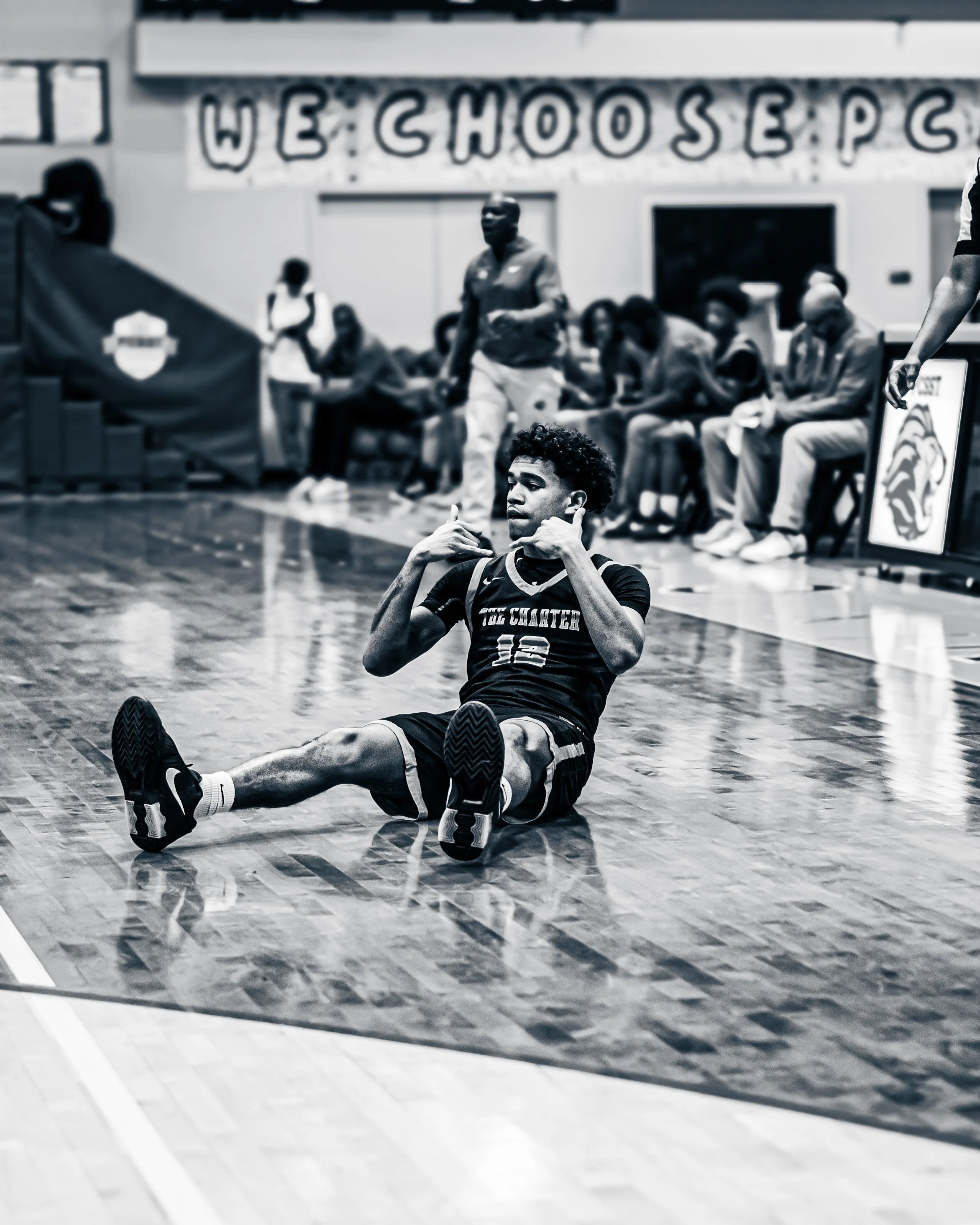 A young male athlete sitting on a gymnasium floor with a focused expression, wearing a black jersey with the words "TAC CHARTER" and the number 12 on it, with his legs extended and hands near his face, in front of a row of seated spectators and a lar