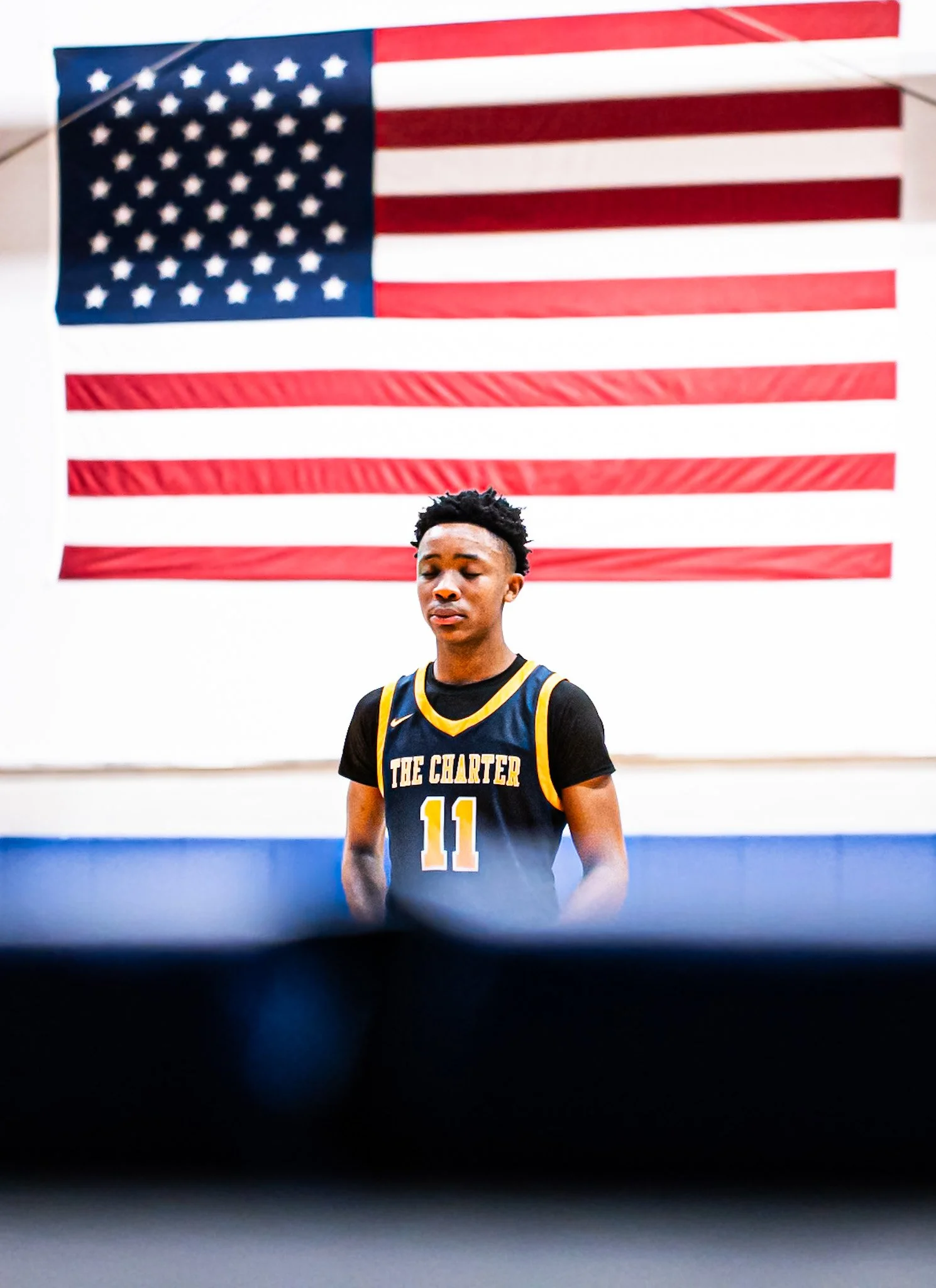 A young man in a basketball uniform with the number 11 and the words "The Charter" on it, standing with eyes closed in front of an American flag.