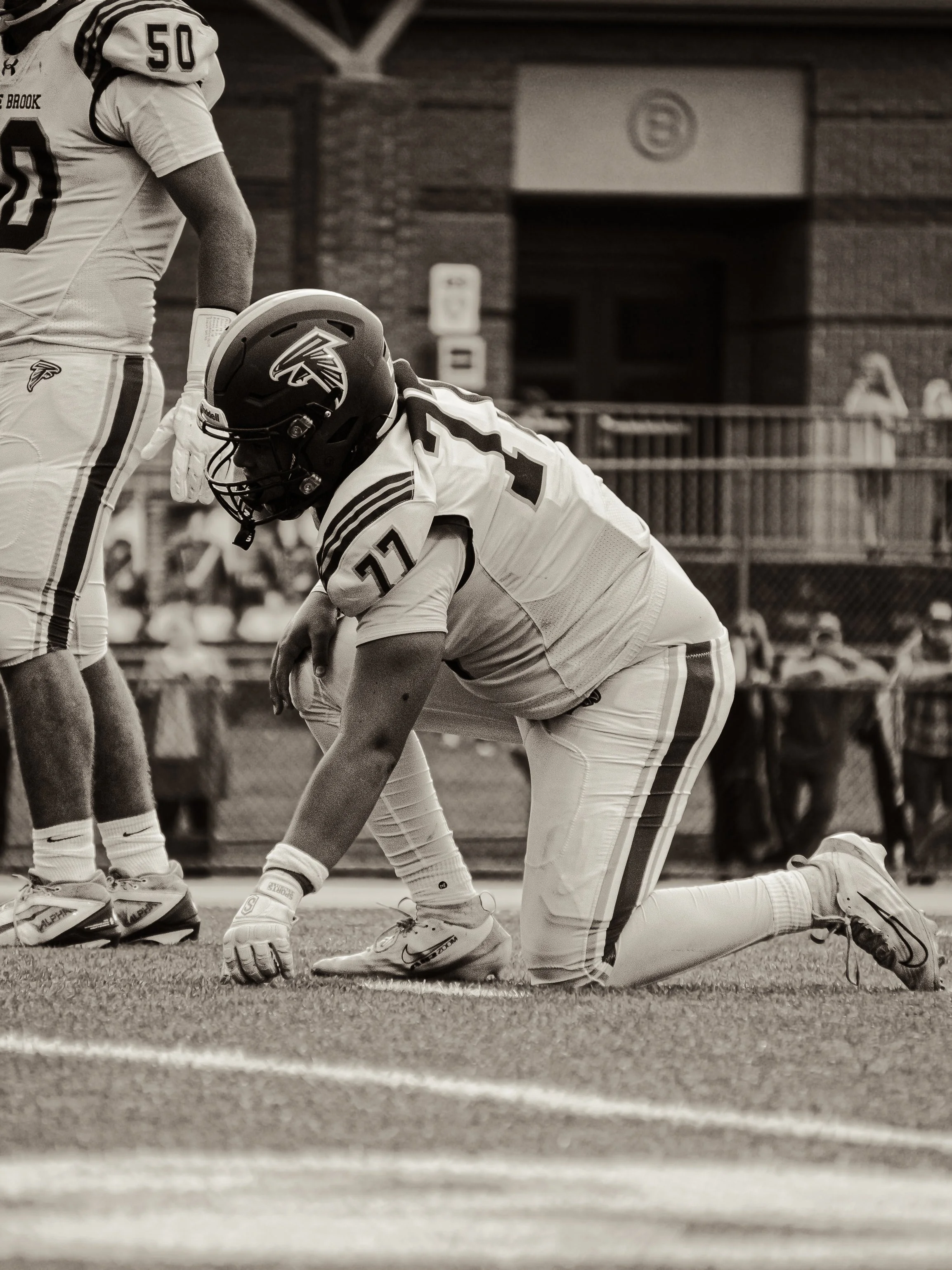 A football player kneeling on the field, wearing helmet and uniform, facing another player standing nearby, with spectators in the background.