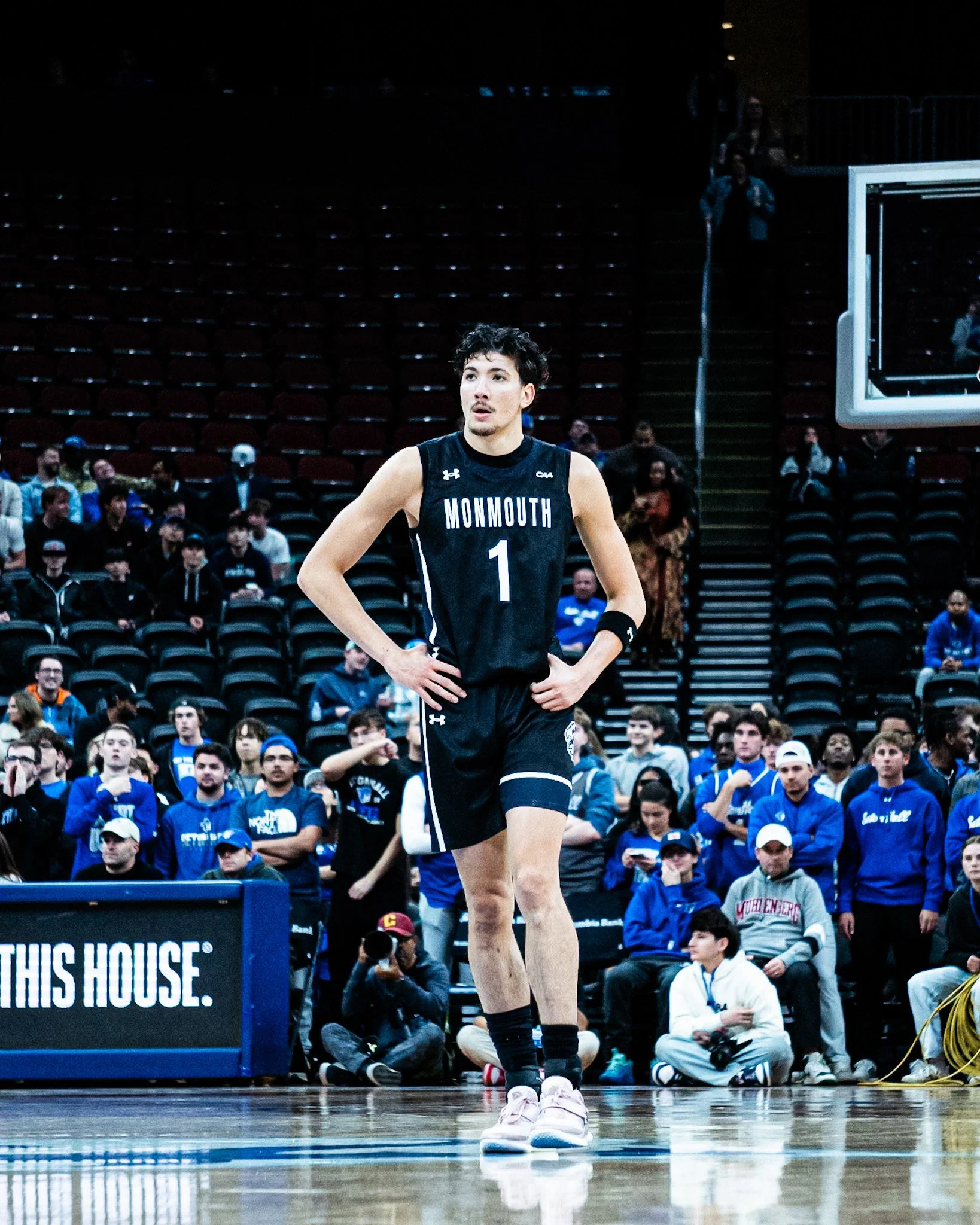 A young male basketball player in a black Monmouth jersey standing on a basketball court with a crowd of spectators in the background.
