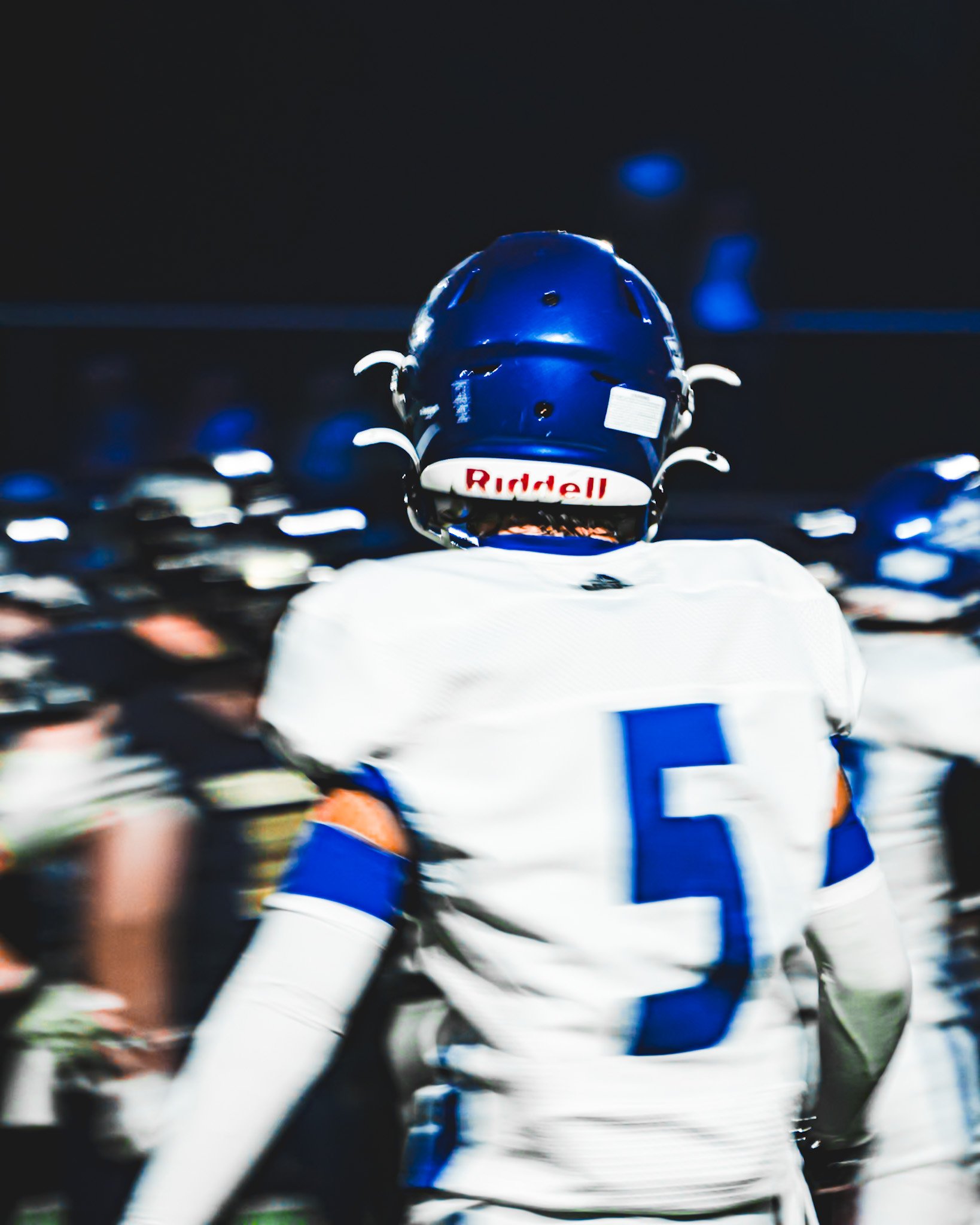 Back of a football player wearing a blue helmet with a white and red logo, white jersey with blue accents, and black gloves, standing among blurred players on the field.