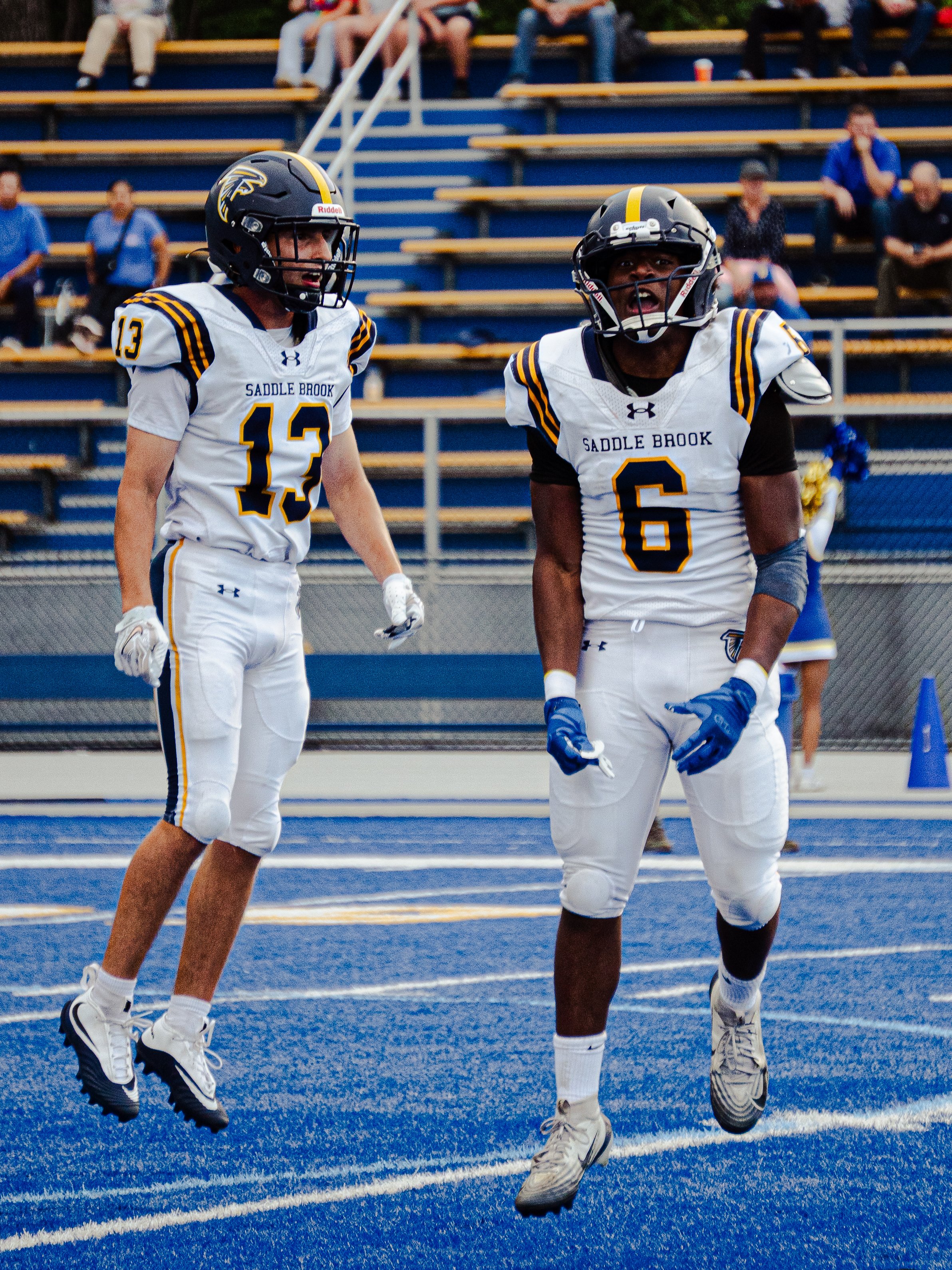 Two boys in football uniforms are on a football field. The boy on the left is wearing a black helmet and white jersey with the number 13, and the boy on the right is also in a helmet and white jersey with the number 6. They are in motion, with the bo