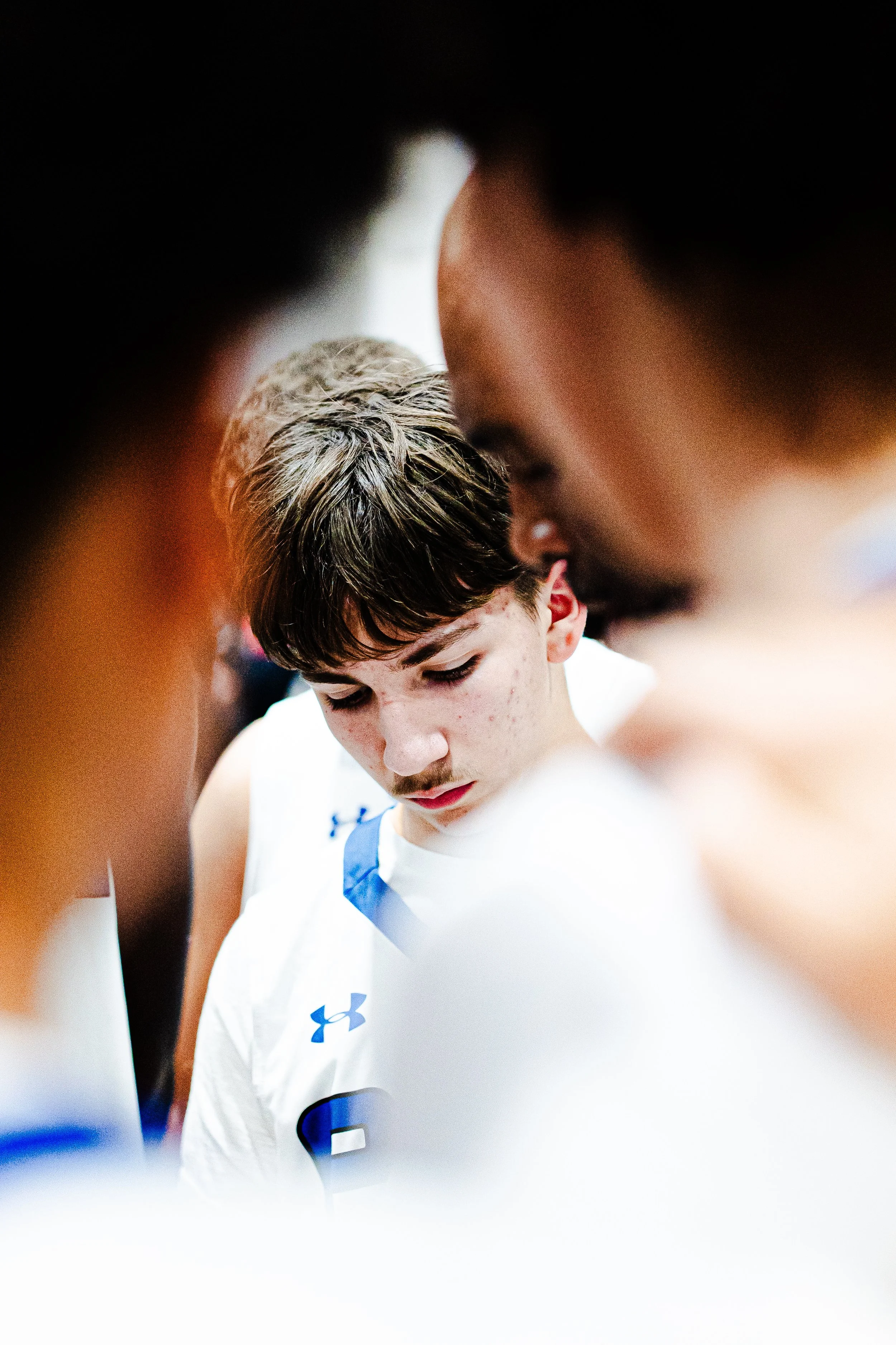 A young male athlete in a white sports jersey with blue accents, possibly a basketball player, is seen looking down during a game or practice.