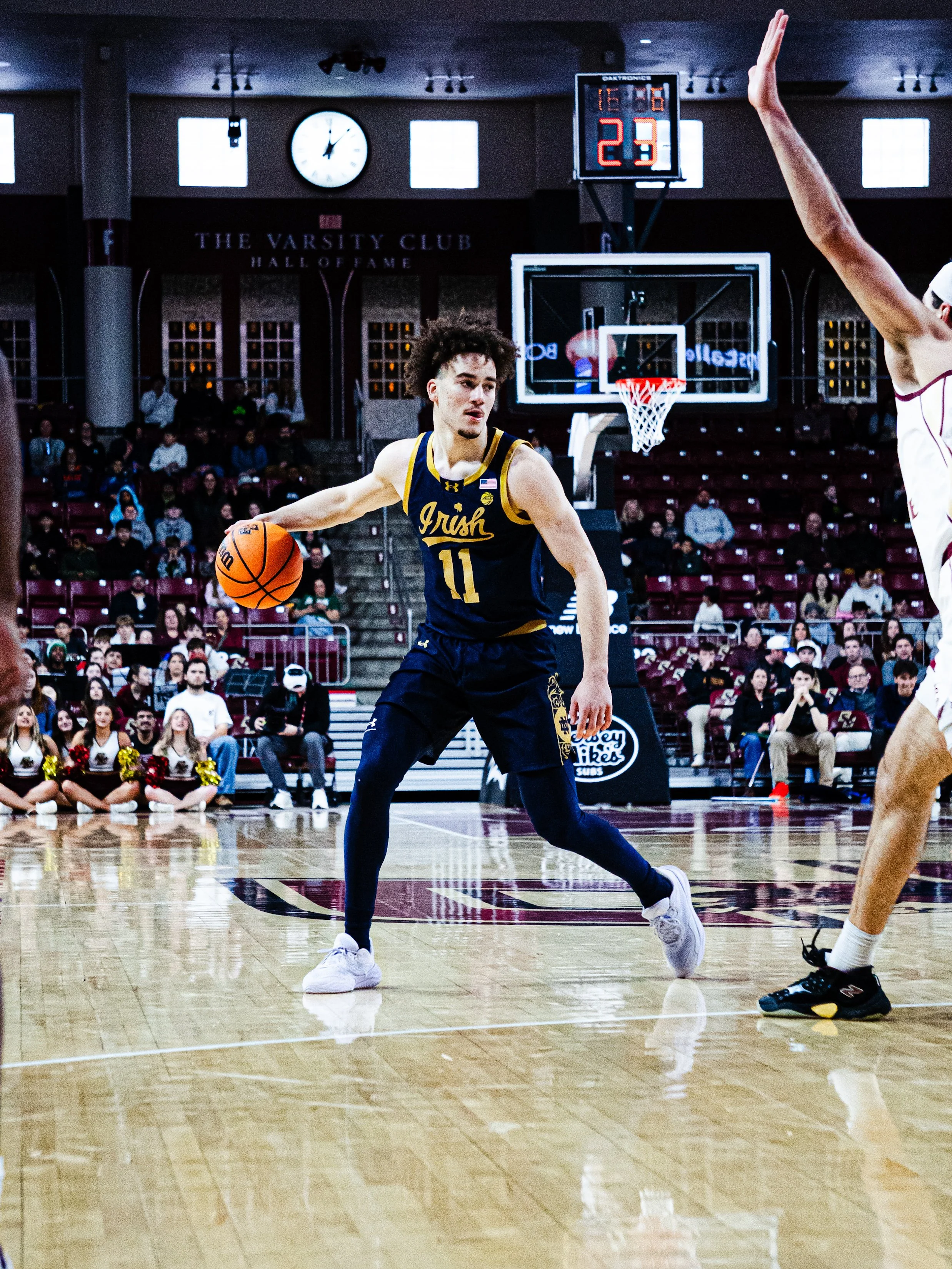 A basketball player wearing a navy and gold jersey with the number 11 dribbling a basketball during a game in an indoor gym. The gym has a scoreboard, a clock, and spectators watching from the bleachers, some wearing cheerleading uniforms.