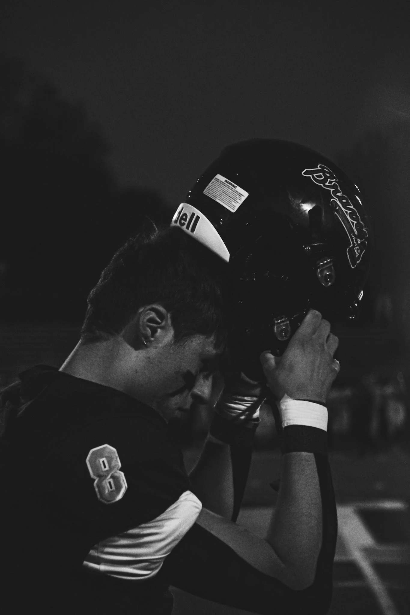 A black and white photo of two men in sports jerseys embracing each other, one with a football helmet. The man in the foreground is wearing a jersey with the number 8.
