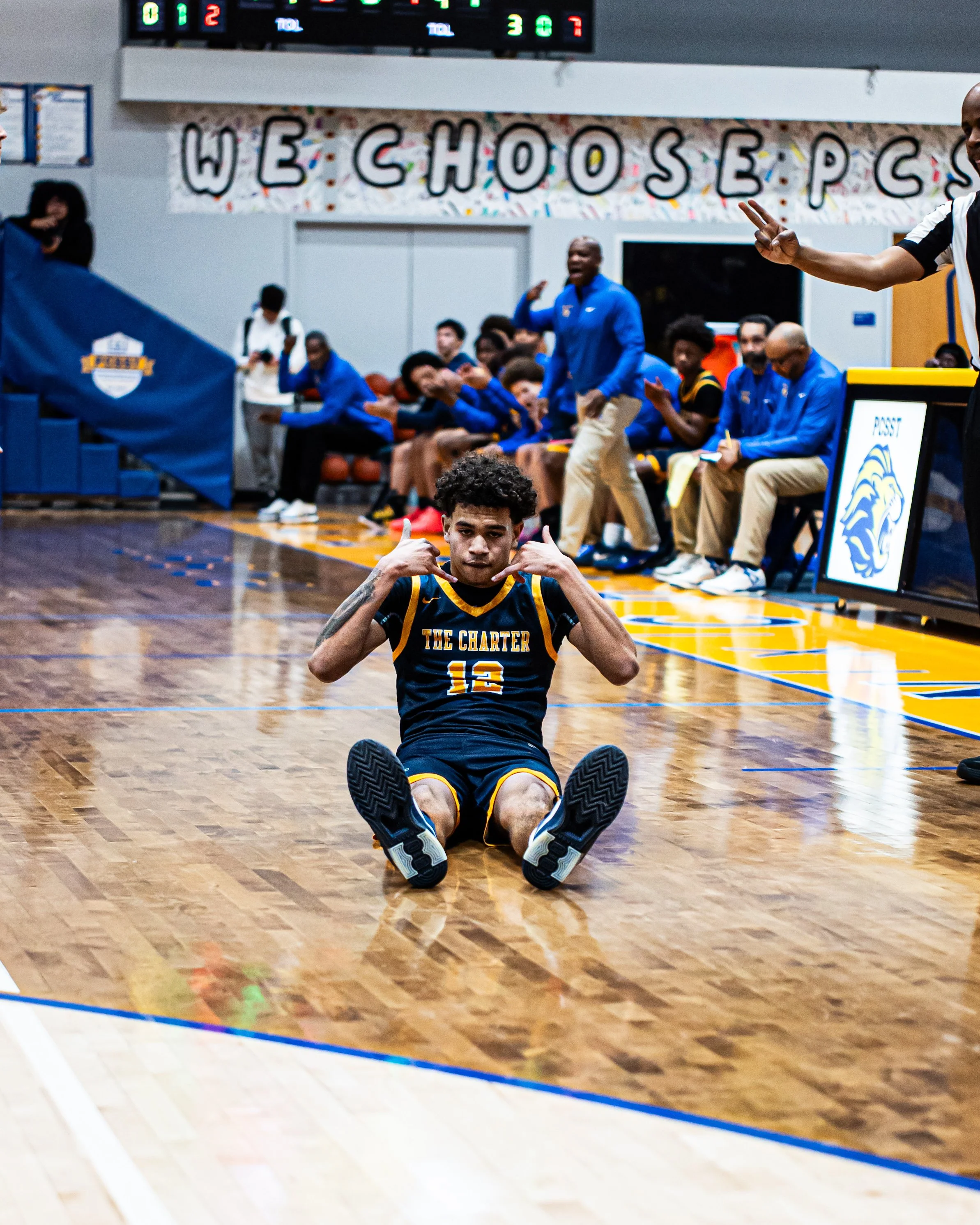 A young man in a basketball uniform sitting on the court floor with his legs stretched out, gesturing with both hands, in a gymnasium with players and coaching staff on the bench behind him and a scoreboard above. There is a colorful banner with the 