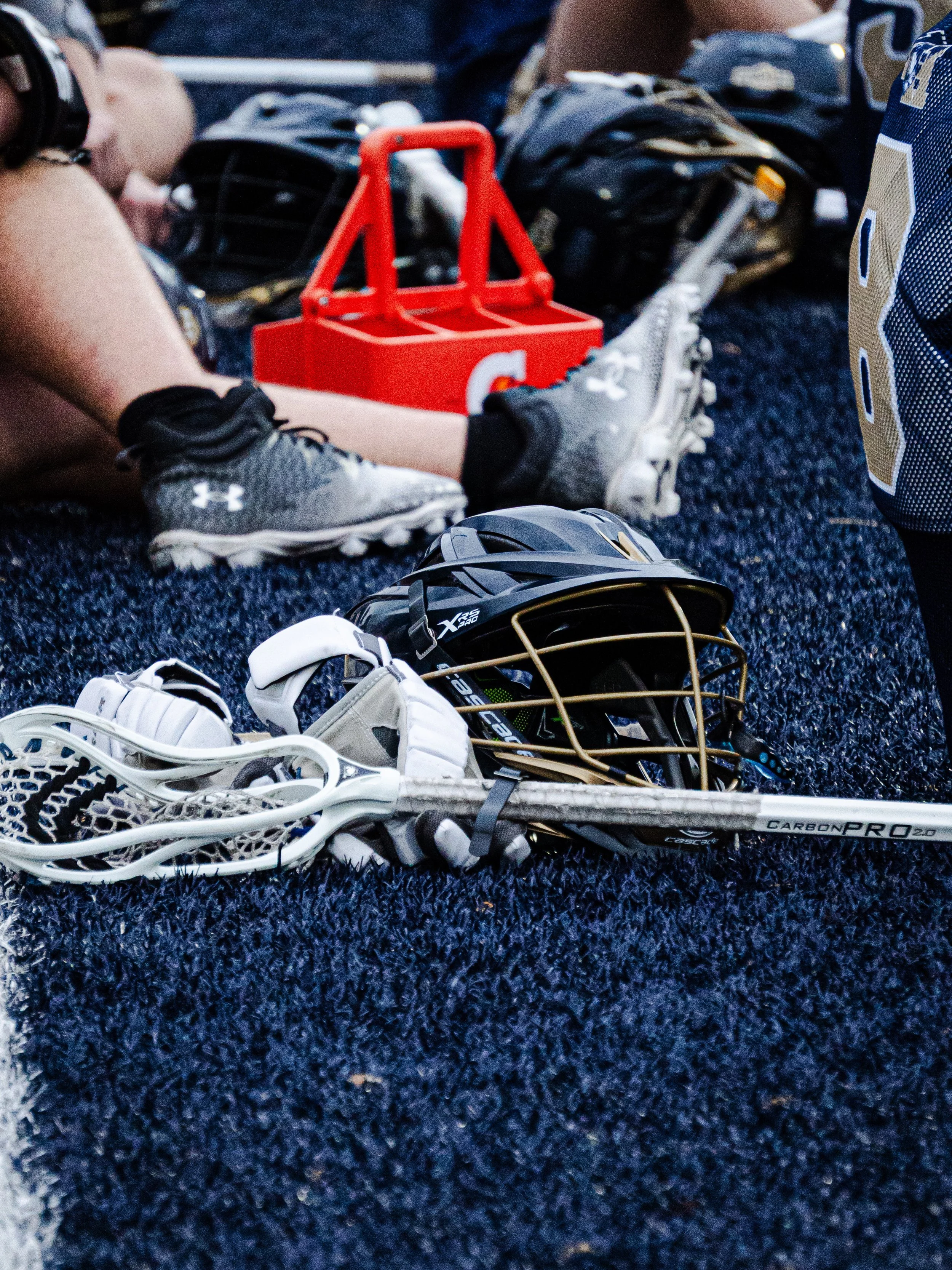 A pile of sports equipment on a dark blue turf field, including a lacrosse stick, a hockey helmet, several pairs of athletic shoes, and a red practice puck used for hockey or street hockey, with a person wearing athletic clothing and a baseball jerse