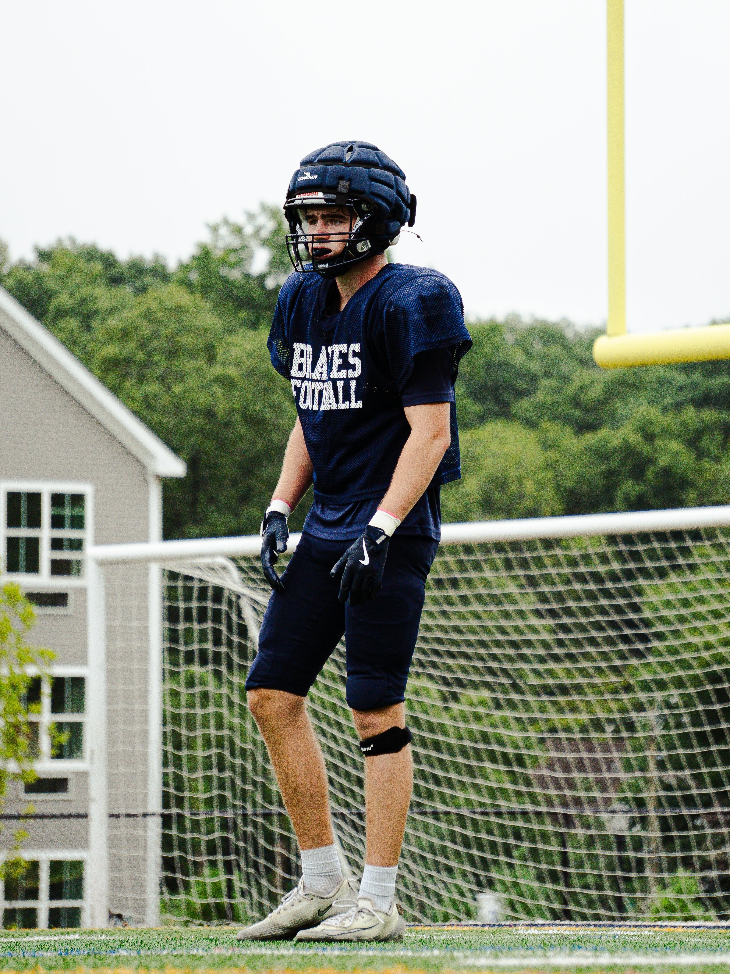 A football player standing on the field wearing a navy blue uniform, helmet, and gloves, near the goal post on a grassy field with trees and a house in the background.