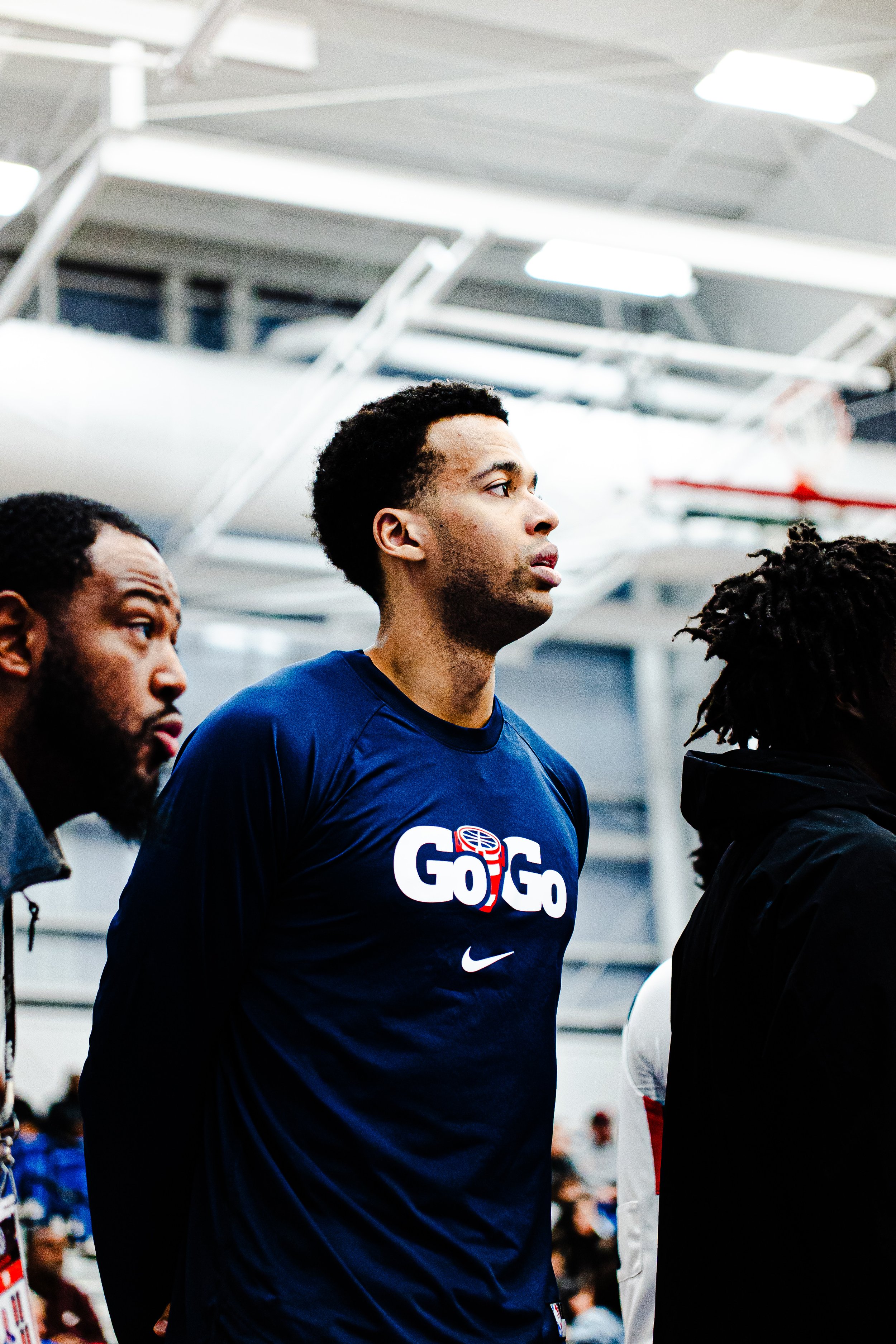 A young man wearing a blue T-shirt with 'G0 G0' logo standing indoors, focused, with other people around.