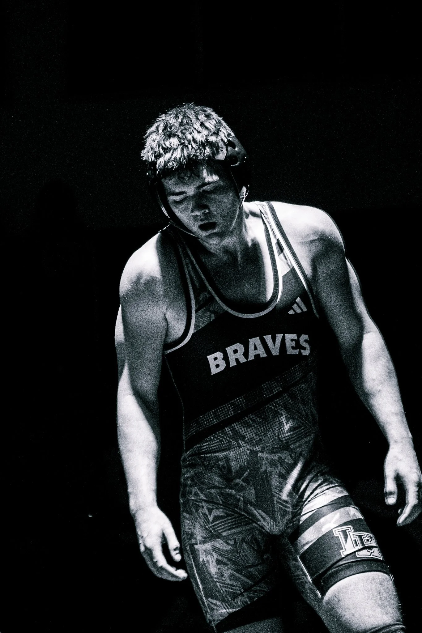 Black and white photo of a male athlete in a sports uniform with the word 'Braves' on his chest, wearing a knee brace and a helmet, with a focused expression, standing against a dark background.