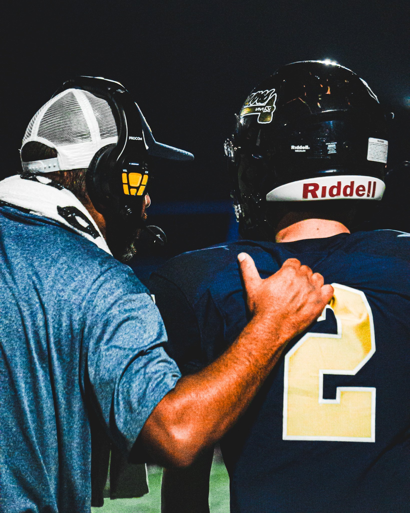 Two football players in uniform are standing closely together on the field, facing away from the camera, with one of them giving a thumbs-up gesture.