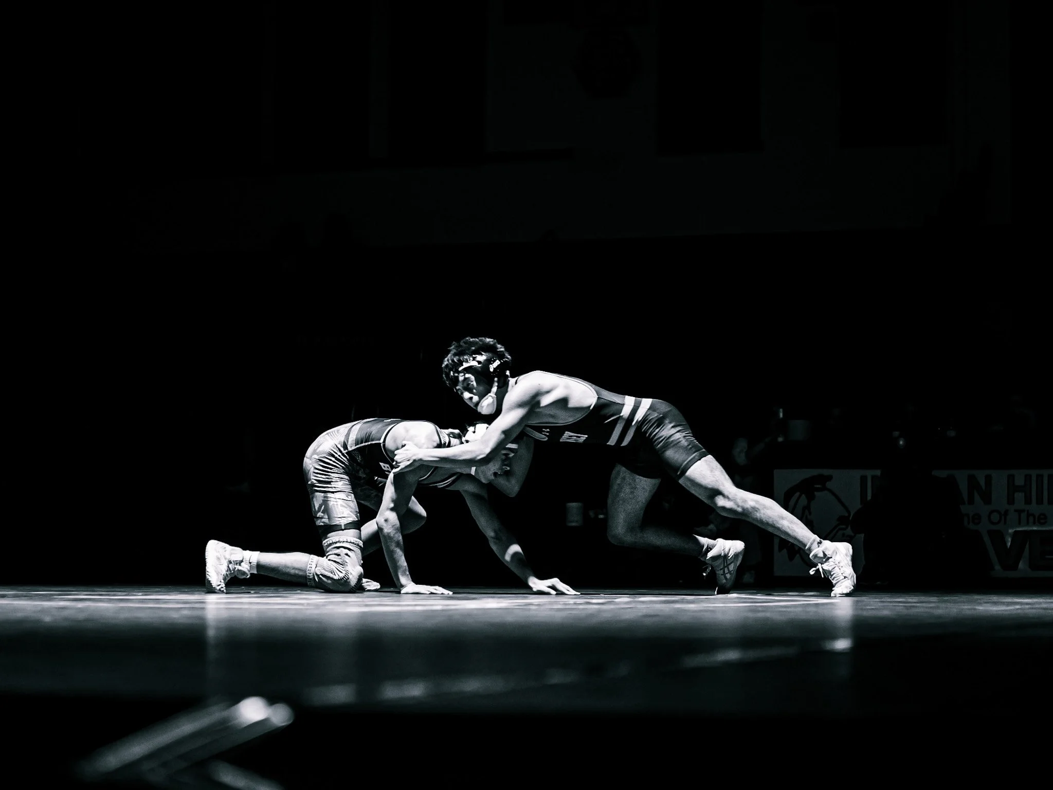 Two wrestlers are engaged in a match on a wrestling mat, with one wrestler on all fours and the other standing and gripping their opponent's arm. The scene is captured in black and white, with a dark background and a spotlight highlighting the wrestl