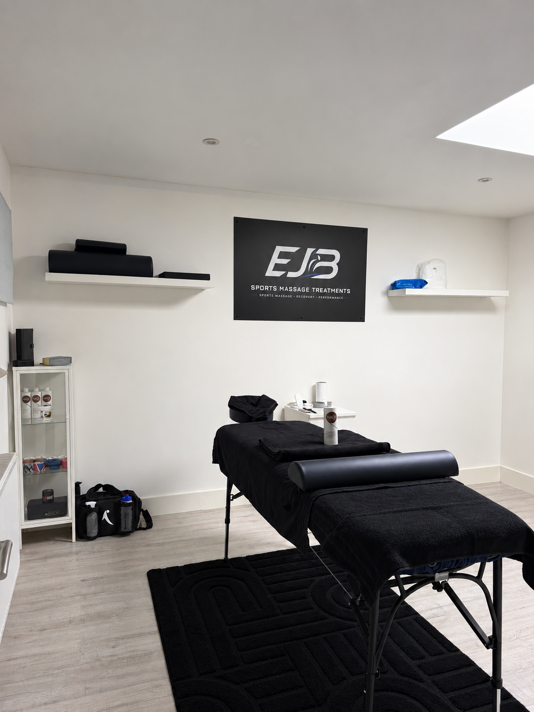 Interior of a sports massage therapy room with a massage table covered in black towels, a black headrest, a white side table with cups, bottles, and tissues, white shelves with black and blue massage equipment, a black area rug, and a black and white sign on the wall reading "EJB Sports Massage Treatments."