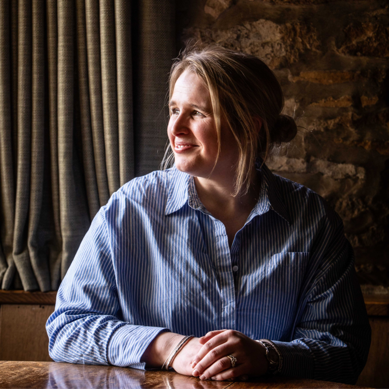 A woman with blonde hair tied back, wearing a blue striped shirt, sitting at a wooden table near a window with curtains, smiling and looking to her left.