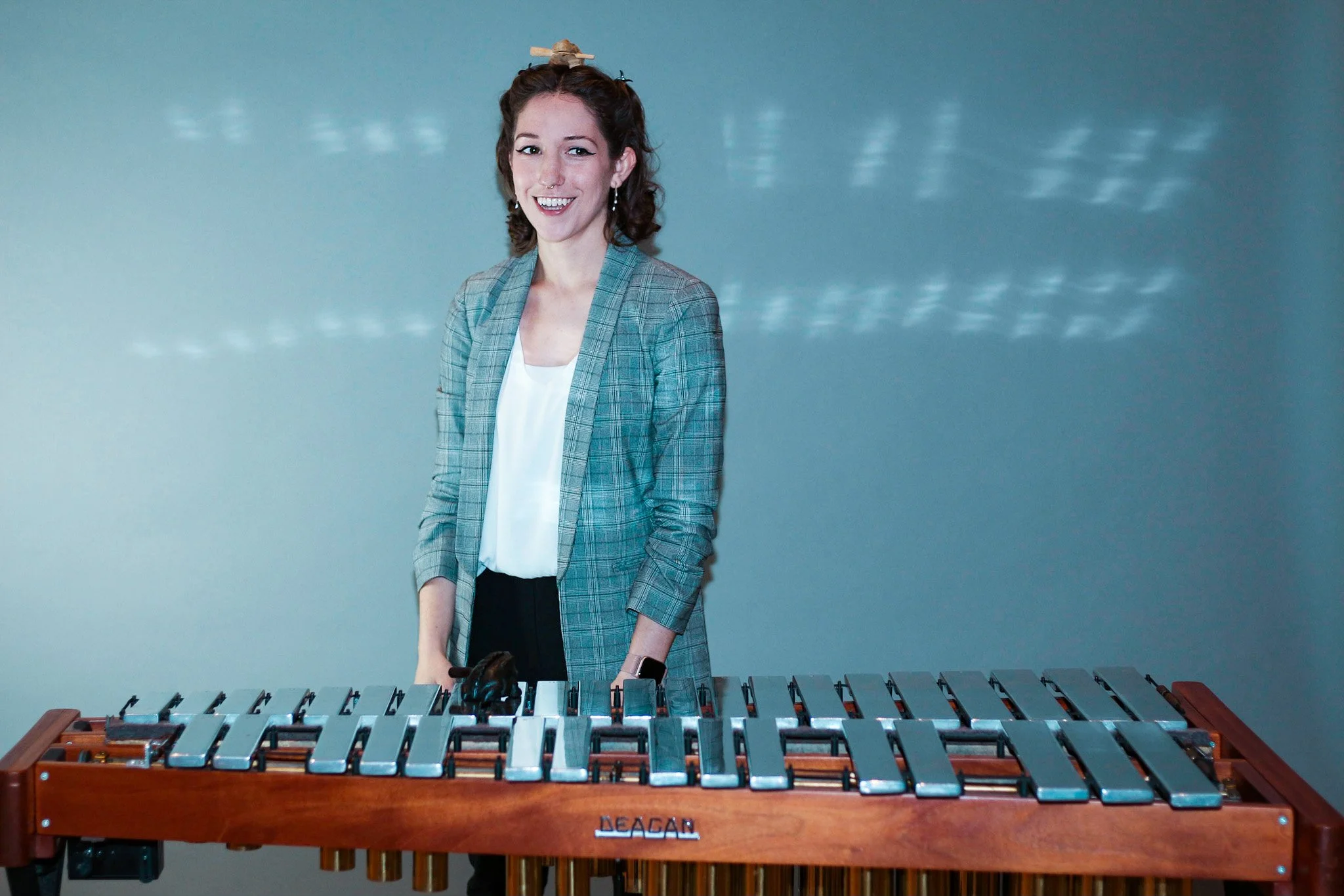 A woman with curly brown hair, wearing a checkered blazer, standing behind a vibraphone, smiling at the camera.