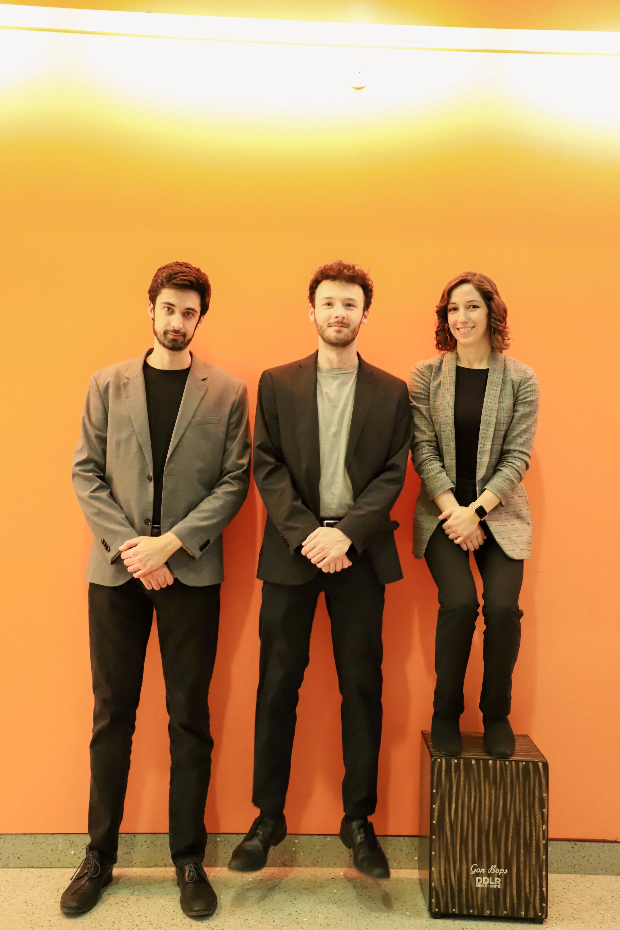 Three young adults standing against a yellow-orange wall, dressed in business casual attire, with a cajón drum on the floor.