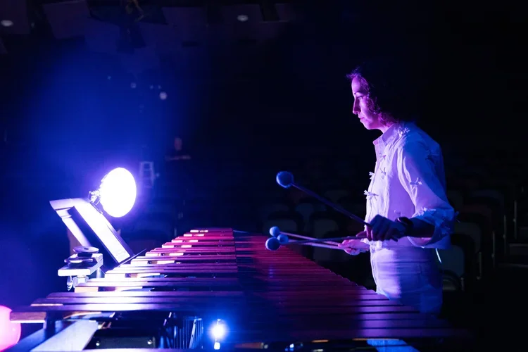 Emma Gierszal performs on a marimba in a dimly lit stage, illuminated by purple and blue lights.