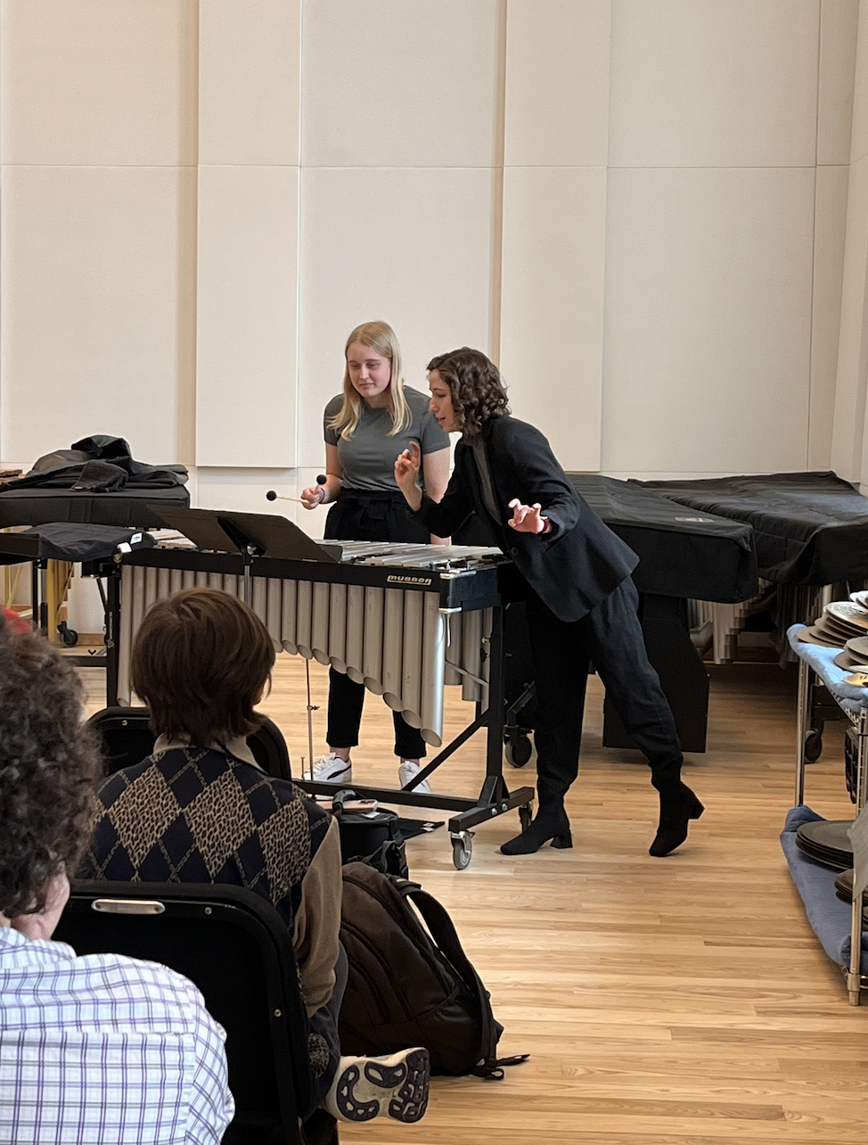 Two women demonstrating a musical instrument to an audience in a room with beige walls and wooden floors.