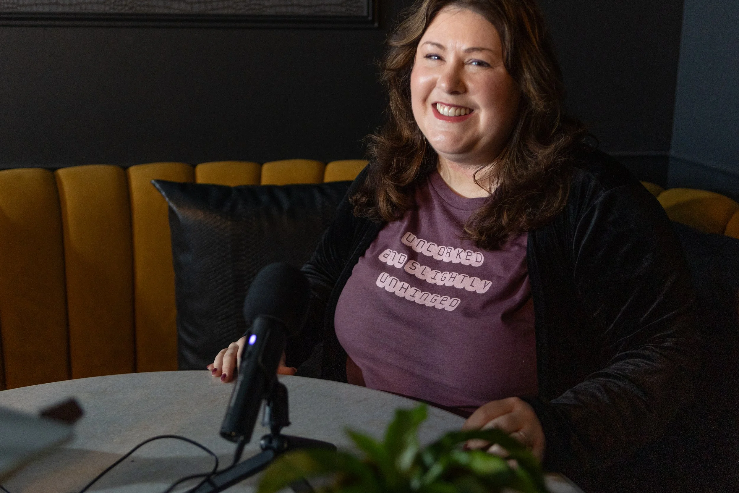 A woman with curly brown hair smiling, sitting at a table, wearing a purple T-shirt and black jacket, with a dark background and yellow chair in the setting.