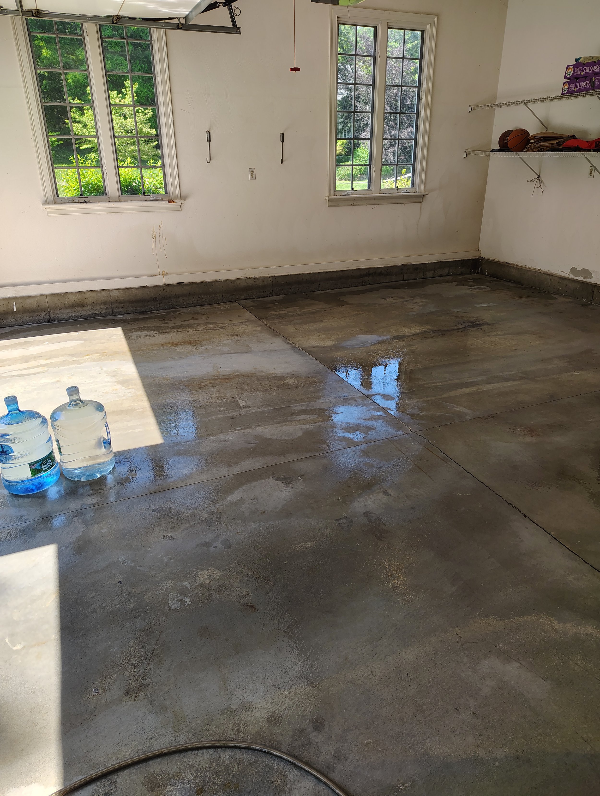 A clean garage with two large windows, a water hose on the floor, and a shelf with a basketball, some boxes, and papers. The floor appears wet, possibly from cleaning or water spill.