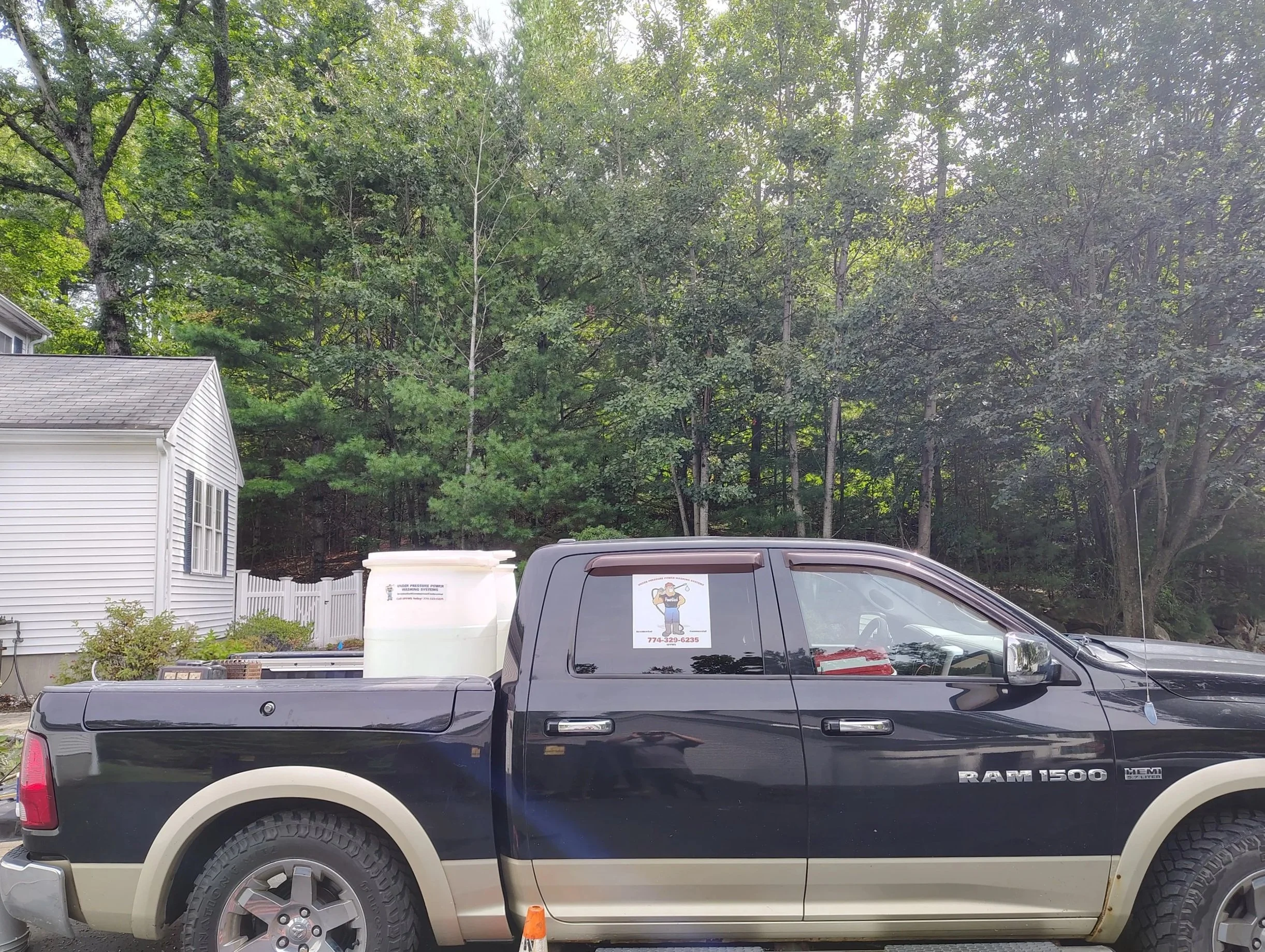 A black RAM 1500 pickup truck parked outdoors in front of a wooded area with green trees. There are orange traffic cones on the ground near the truck and a white house with a gray roof to the left.