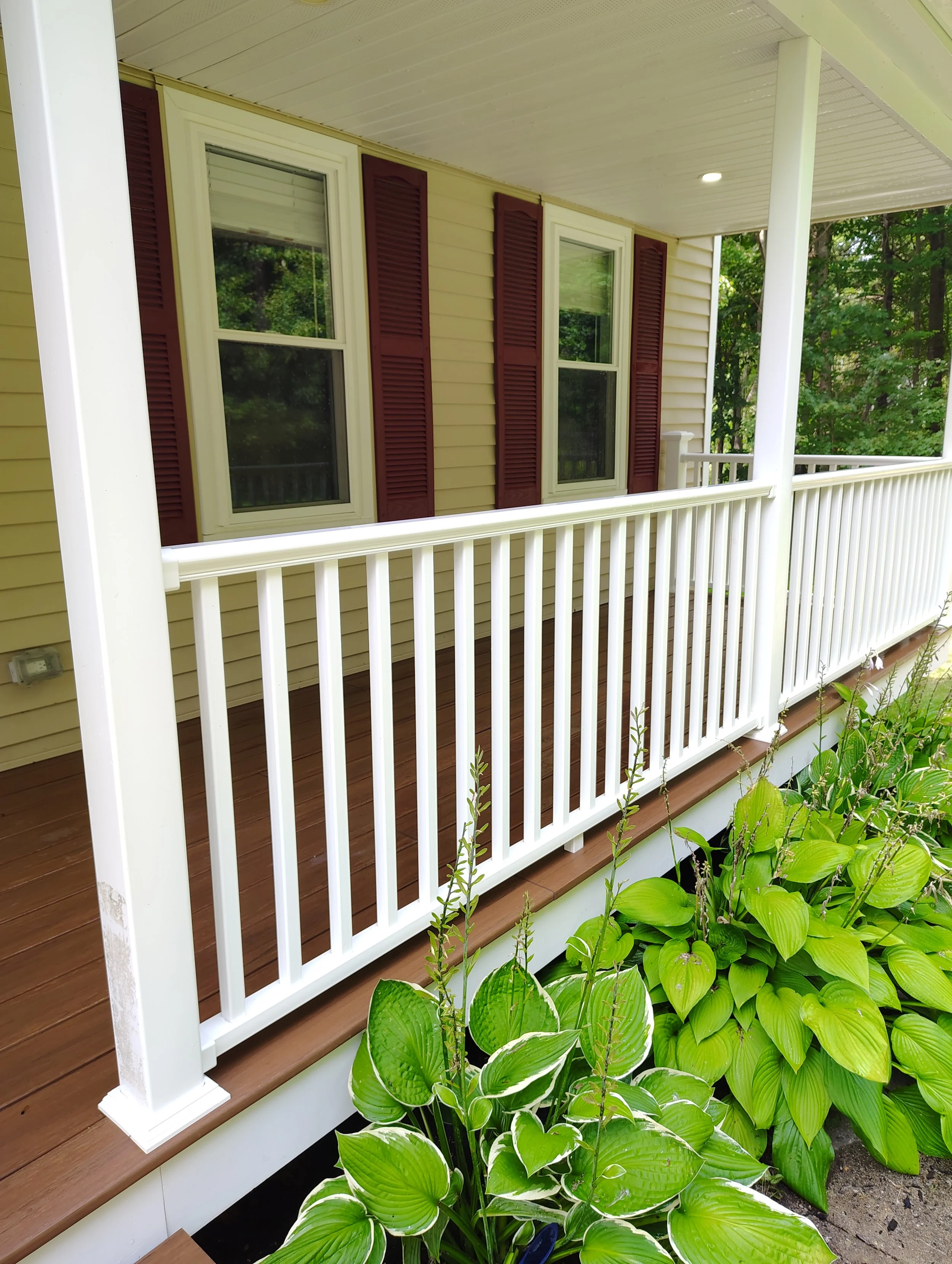 A porch with a white railing and posts, a brown wooden floor, beige siding, two windows with white frames and closed blinds, surrounded by green plants with large leaves.