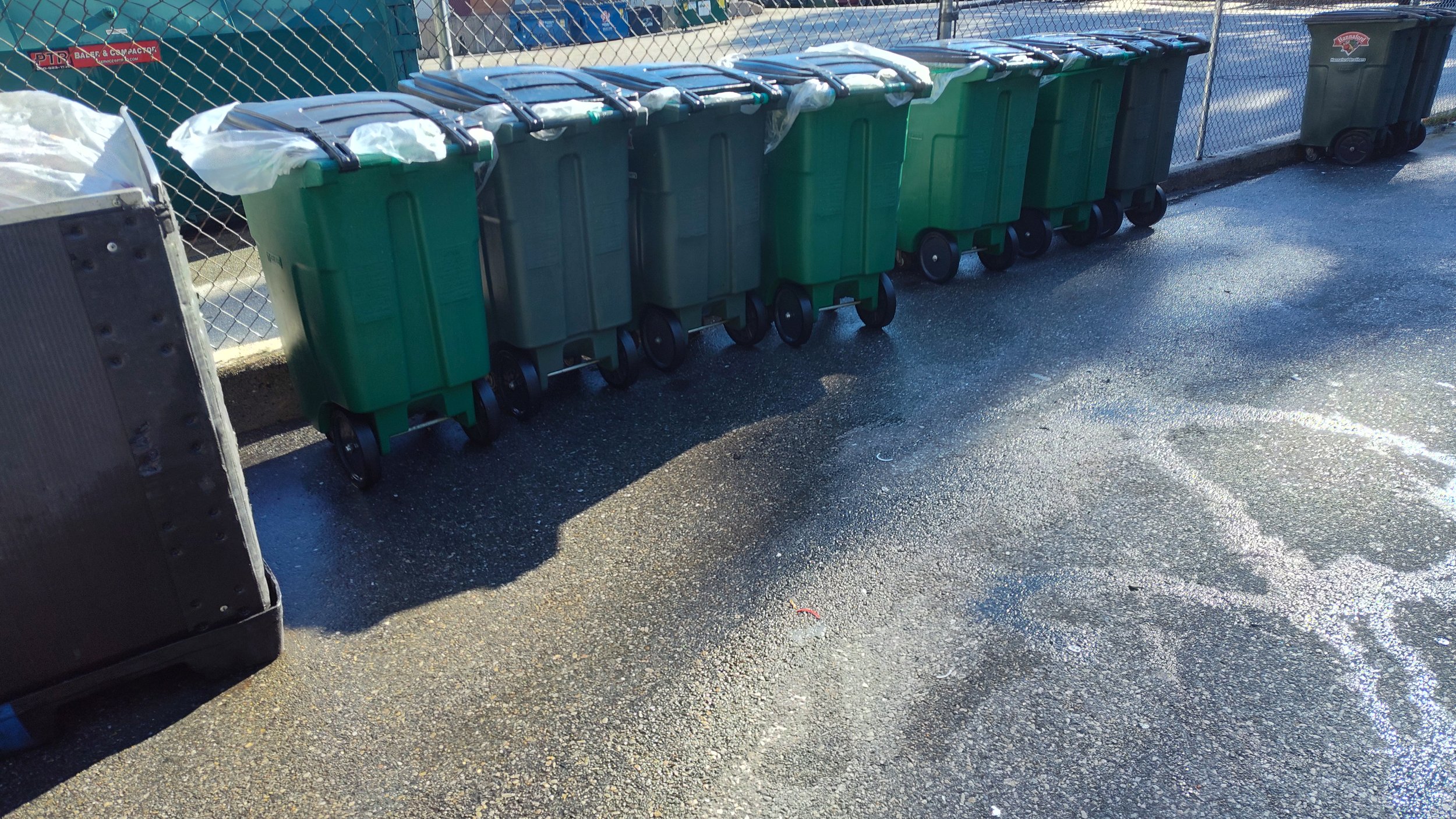 A row of green and black waste and recycling trash bins placed along a chain-link fence, on a wet asphalt surface with sunlight casting shadows.