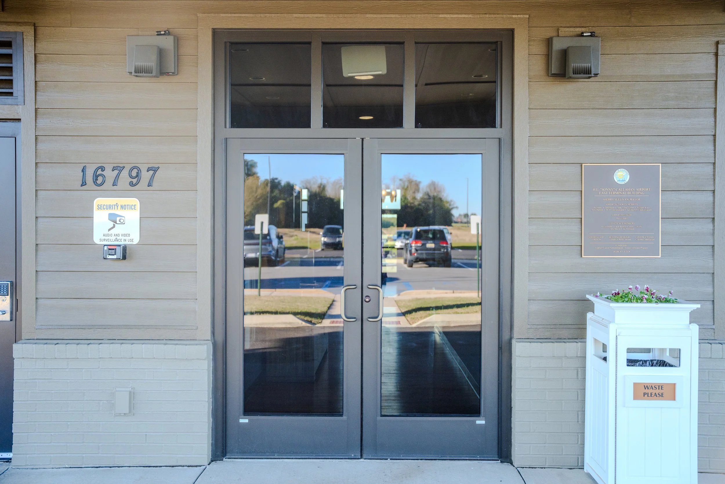 Glass double doors at the entrance of a building with the address 16797 on the wall to the left. There is a security notice sign and a waste bin with a sign that says "Waste Please" on the right side. The reflection in the glass shows parked cars and