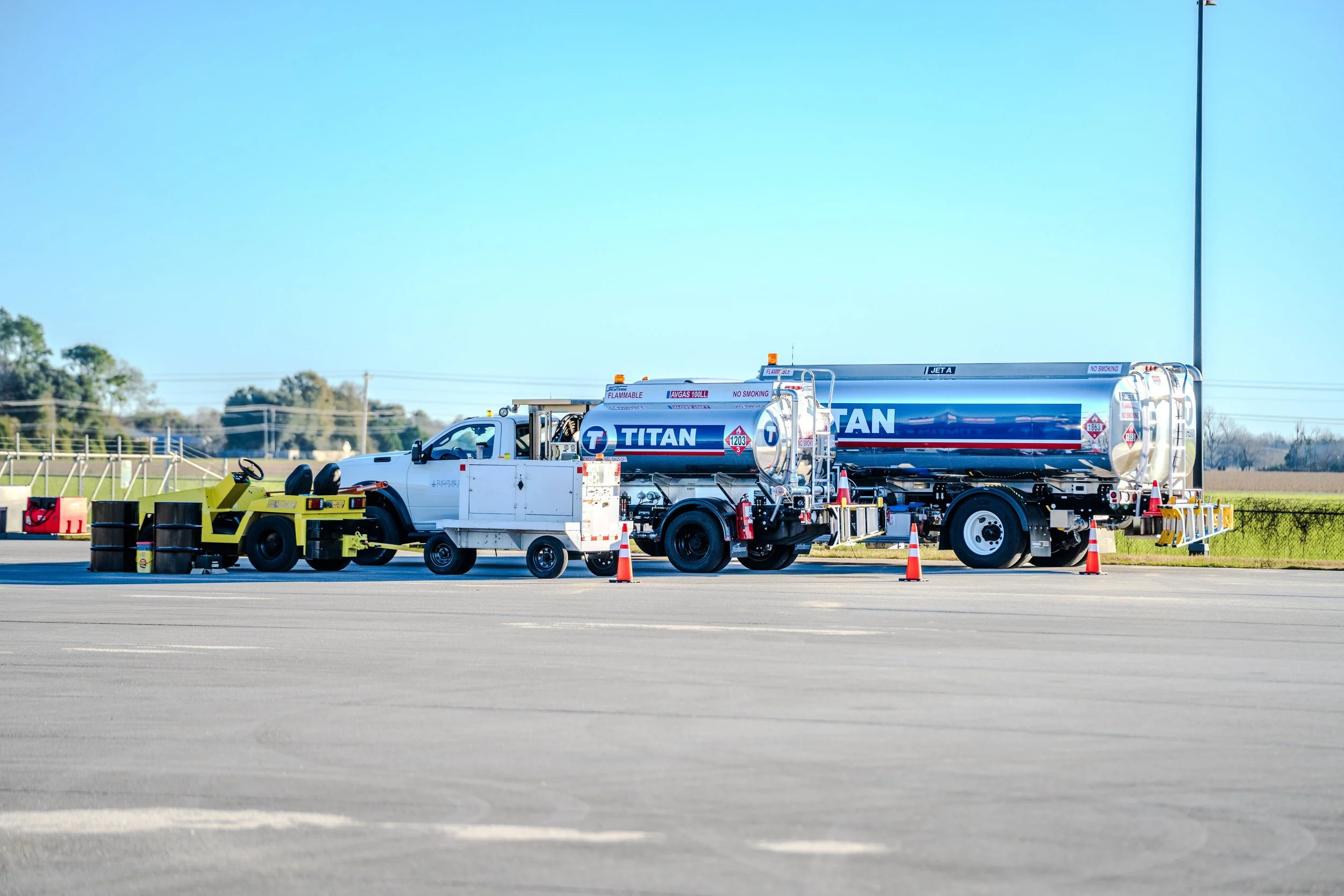 Refueling or loading a large Titan fuel tanker truck at an airport or industrial area, with safety cones and equipment nearby under clear sky.