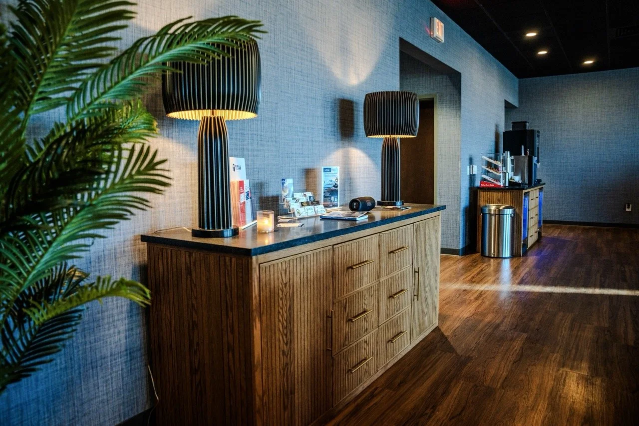 Interior of a room with a wooden sideboard, two black striped lamps, brochures, a candle, and a speaker on top. A large potted plant is partially visible on the left, with blue walls, wooden flooring, and an exit sign above a door.