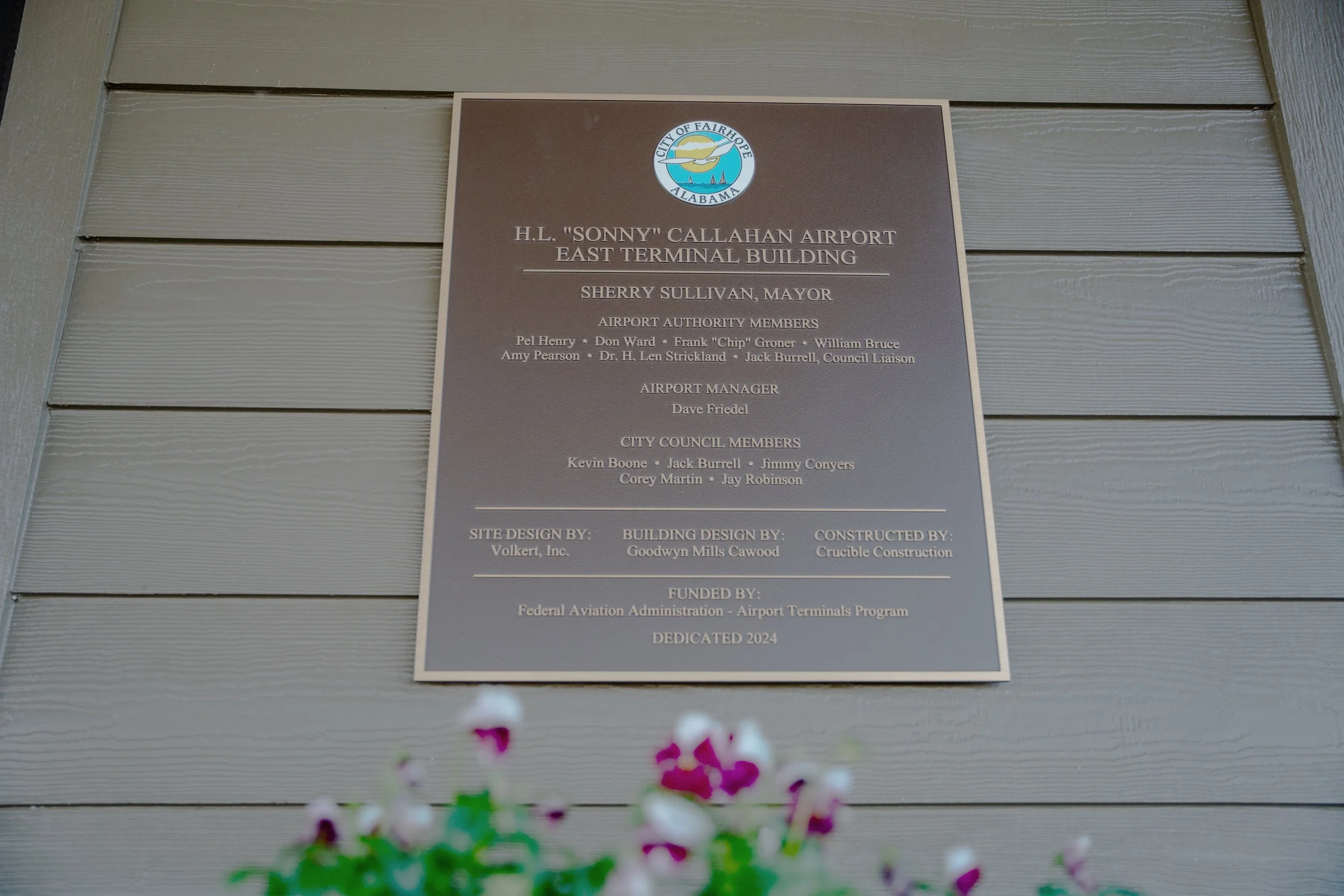 A sign on a building wall reads: H.L. "Sonny" Callahan Airport East Terminal Building, with details of city officials, project designers, and funding sources underneath. The wall consists of horizontal gray wooden planks.